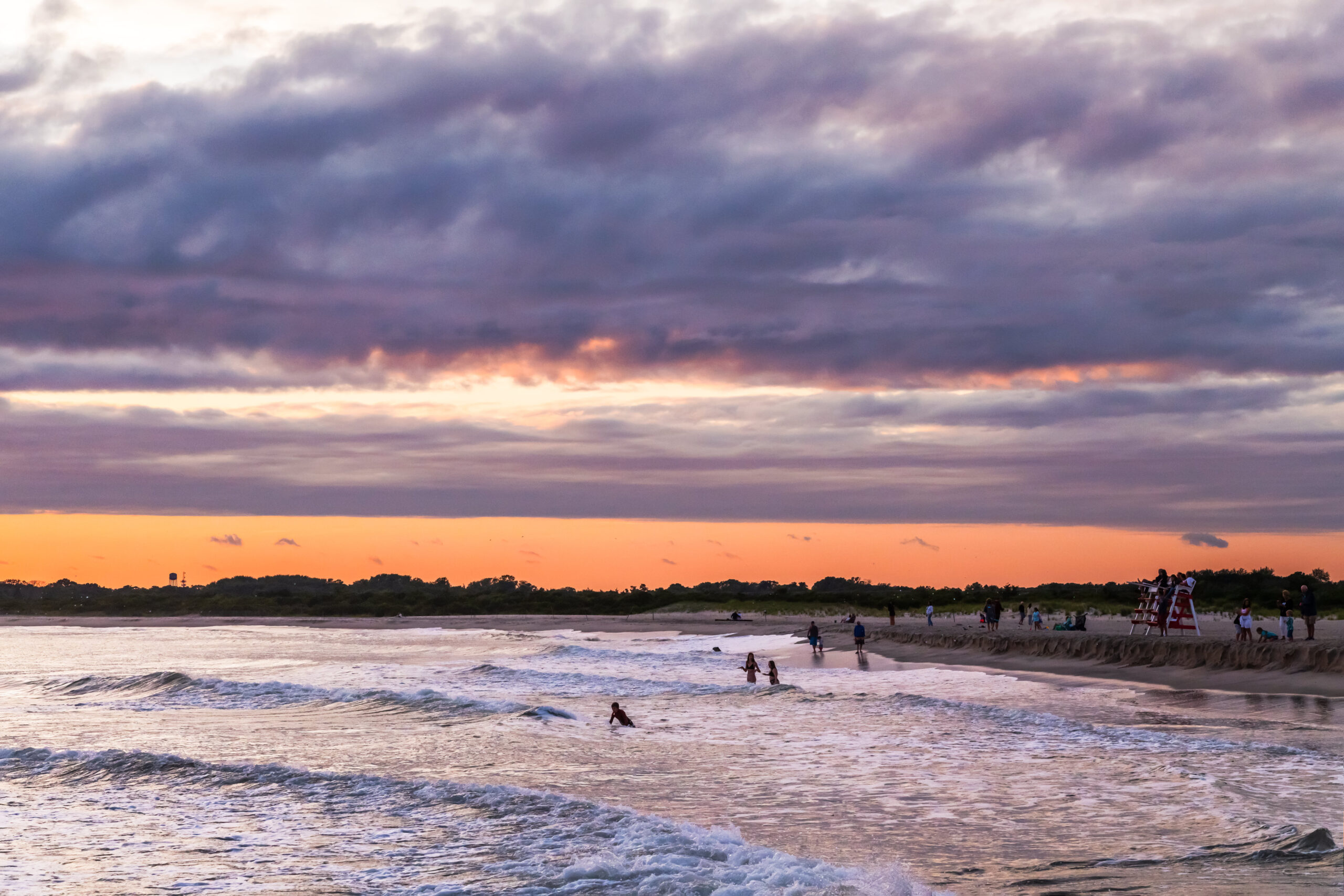 People going in the ocean and sitting on a lifeguard stand at sunset with purple and pink clouds in the sky