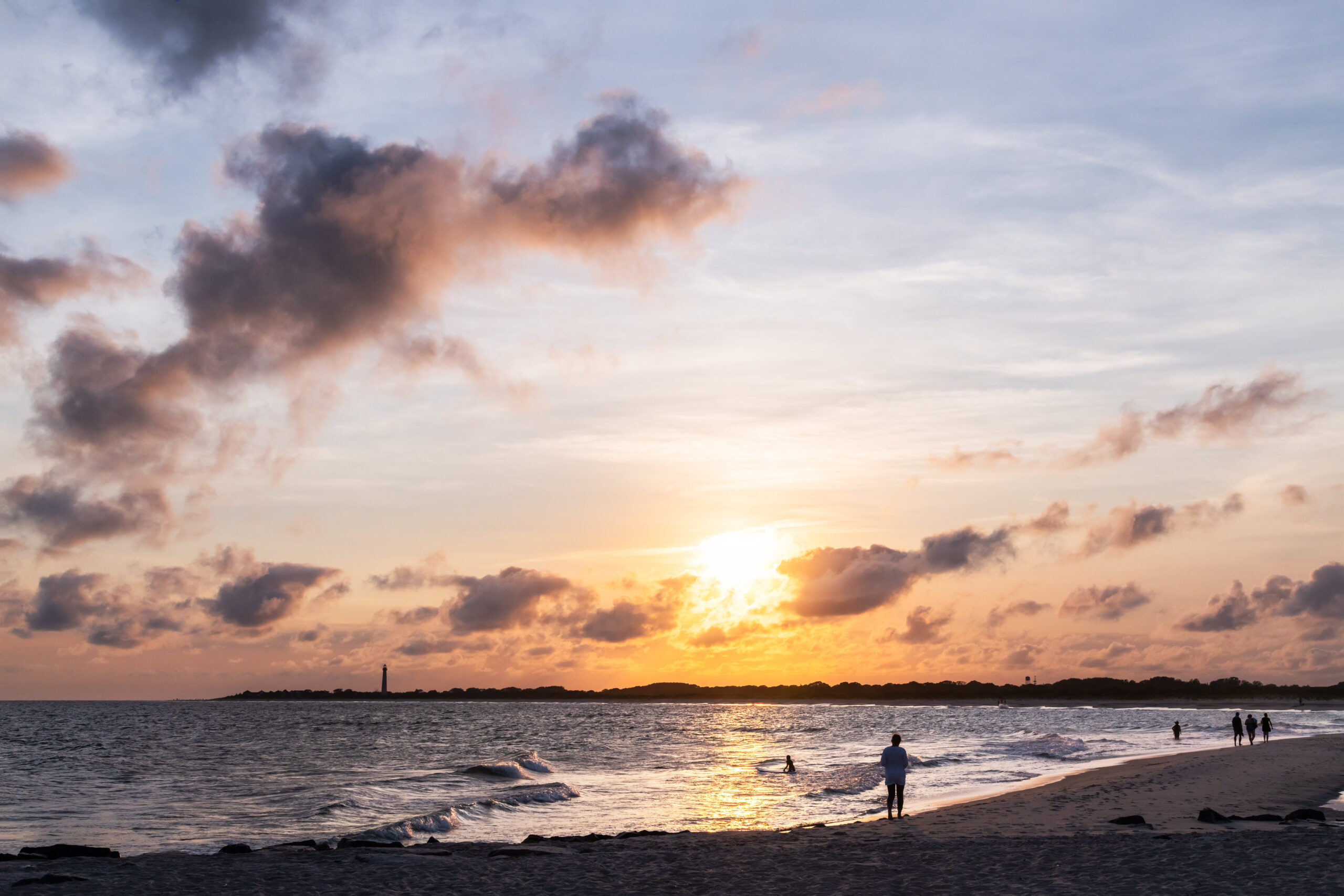 People walking along the ocean at sunset with a few puffy pink and purple clouds in the sky