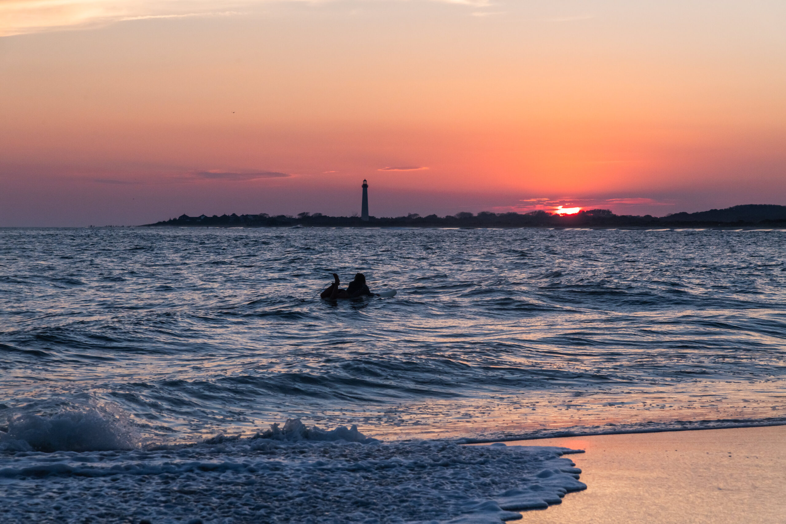 A person on a surfboard in the ocean watching the sunset by the Cape May Lighthouse with waves coming into the shore