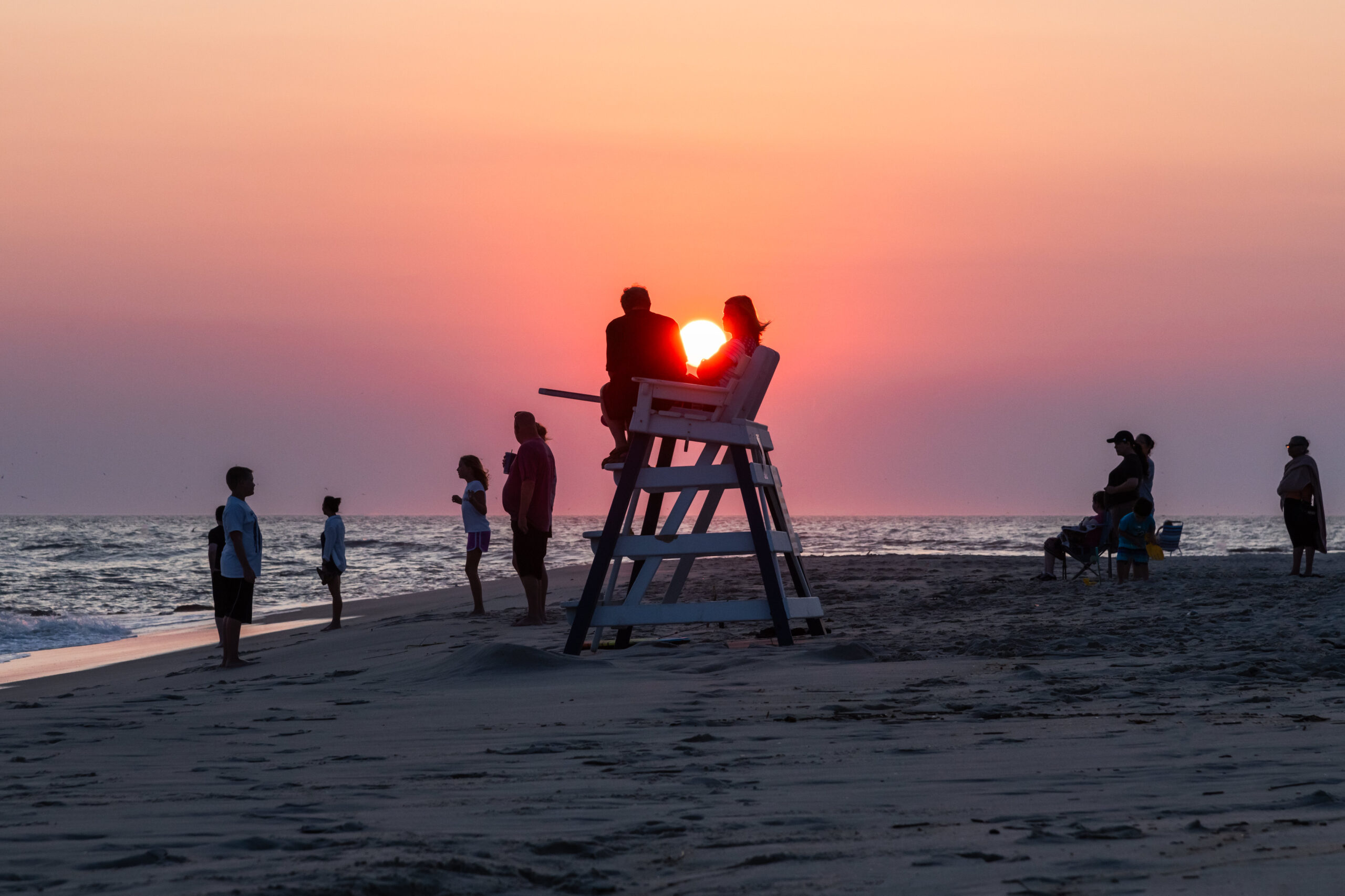 The sun setting behind people sitting on a lifeguard stand and people standing at the ocean