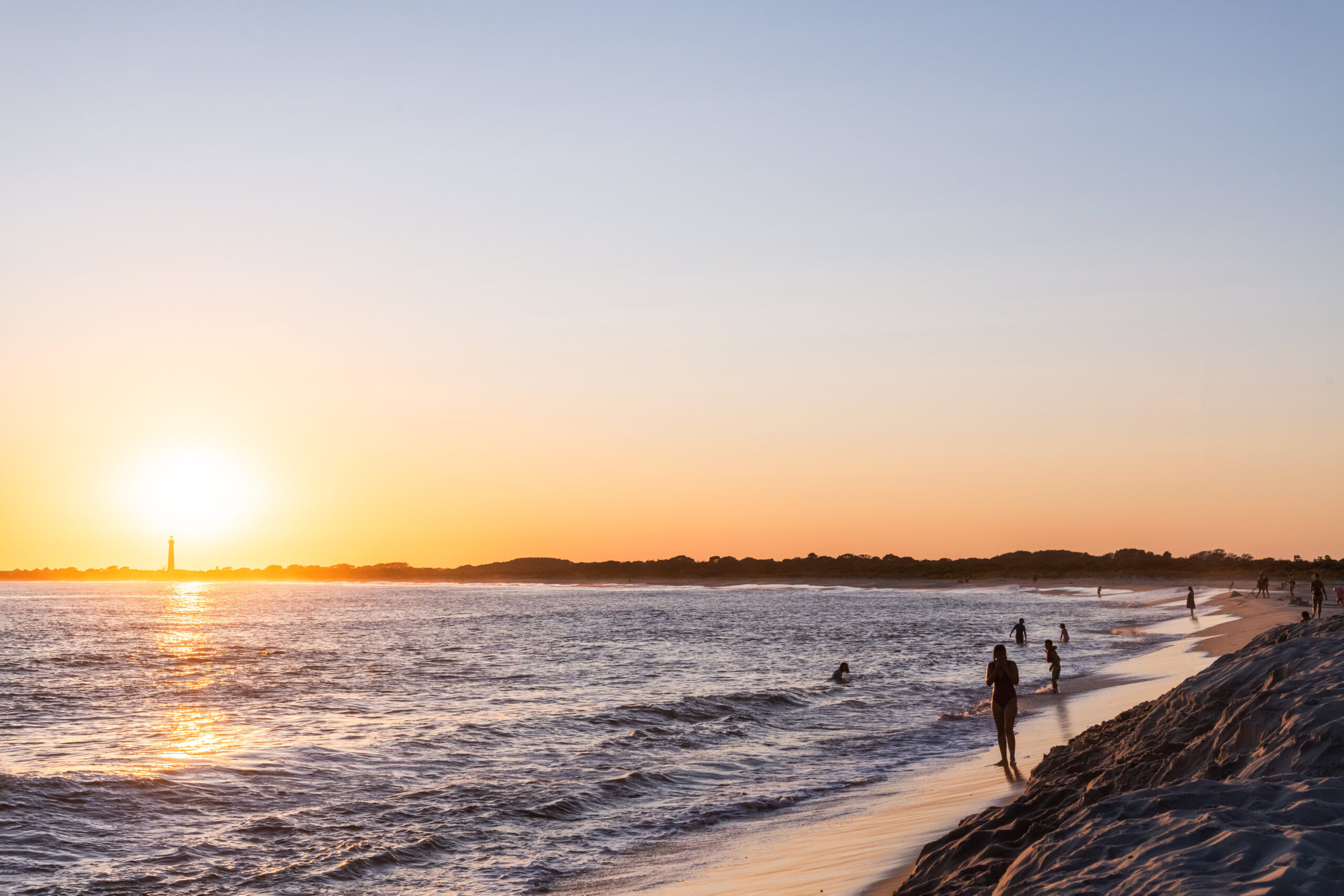 The sun setting in a clear sky with people dipping into the ocean and the Cape May Lighthouse in the distance