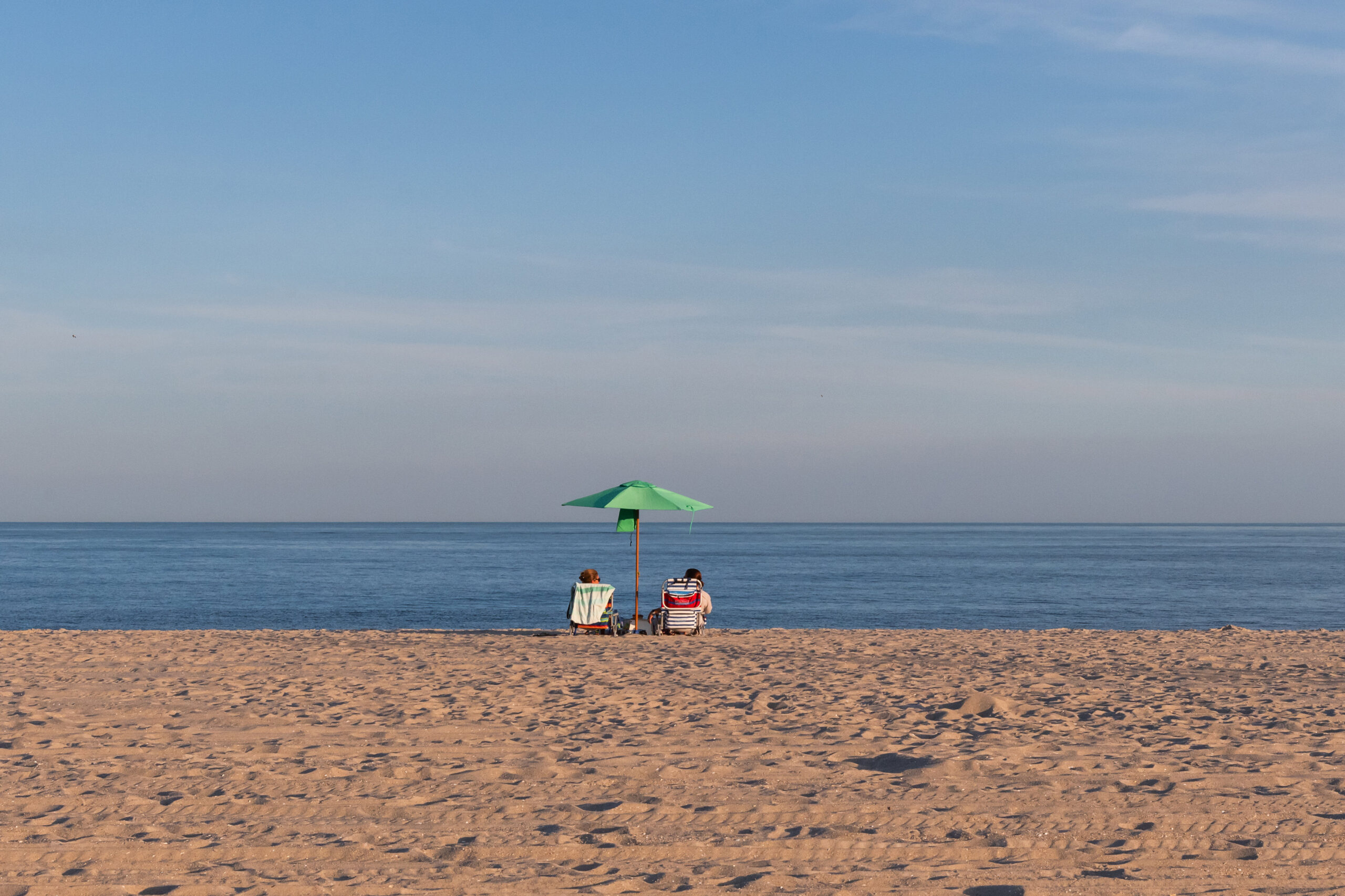 Two people sitting under a green beach umbrella on a clear day with a blue sky