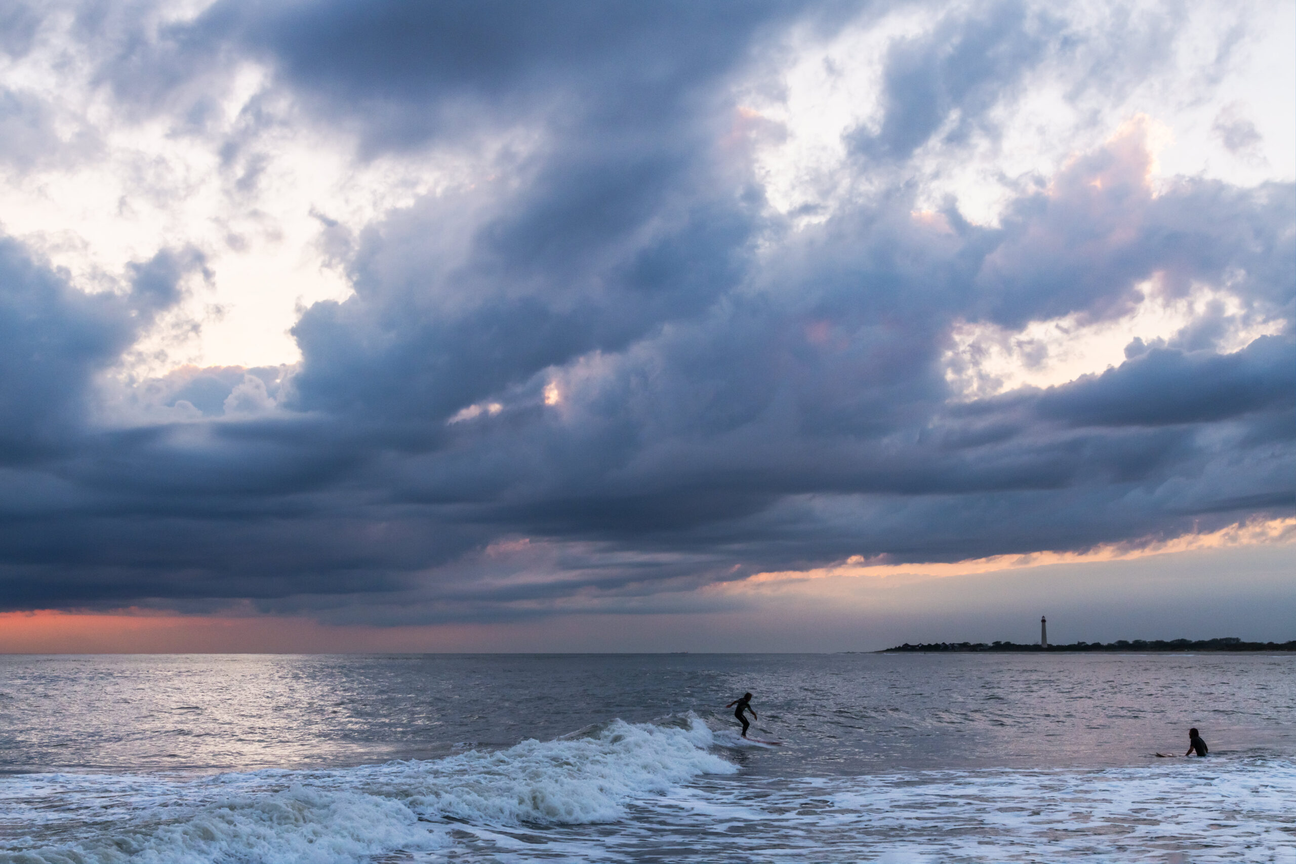 Blue puffy clouds in the sky at sunset with the Cape May Lighthouse in the distance, some pink sky at the horizon, and two people surfing in the ocean
