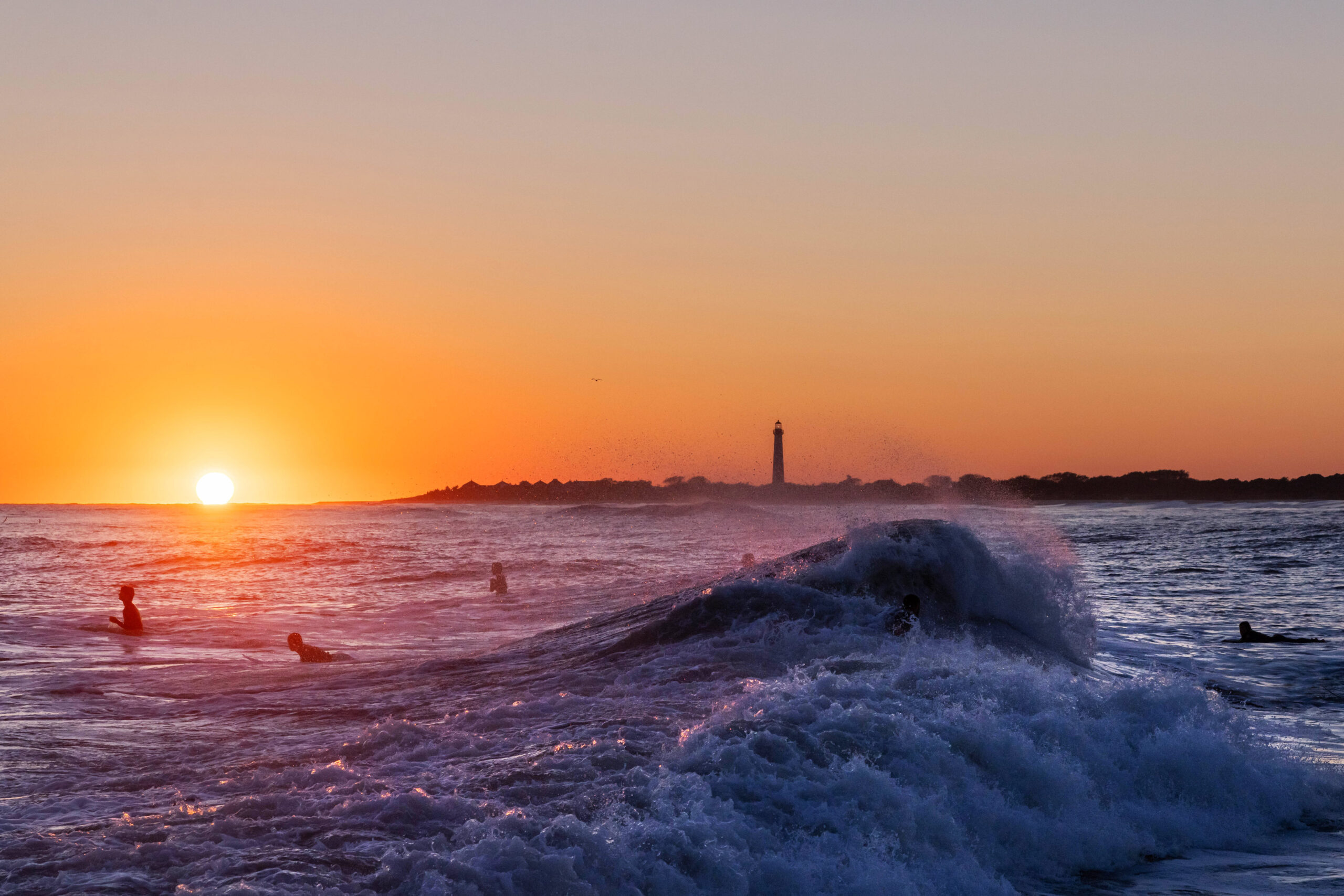 A big wave crashing with surfers in the ocean, and the Cape May Lighthouse in the distance at sunset