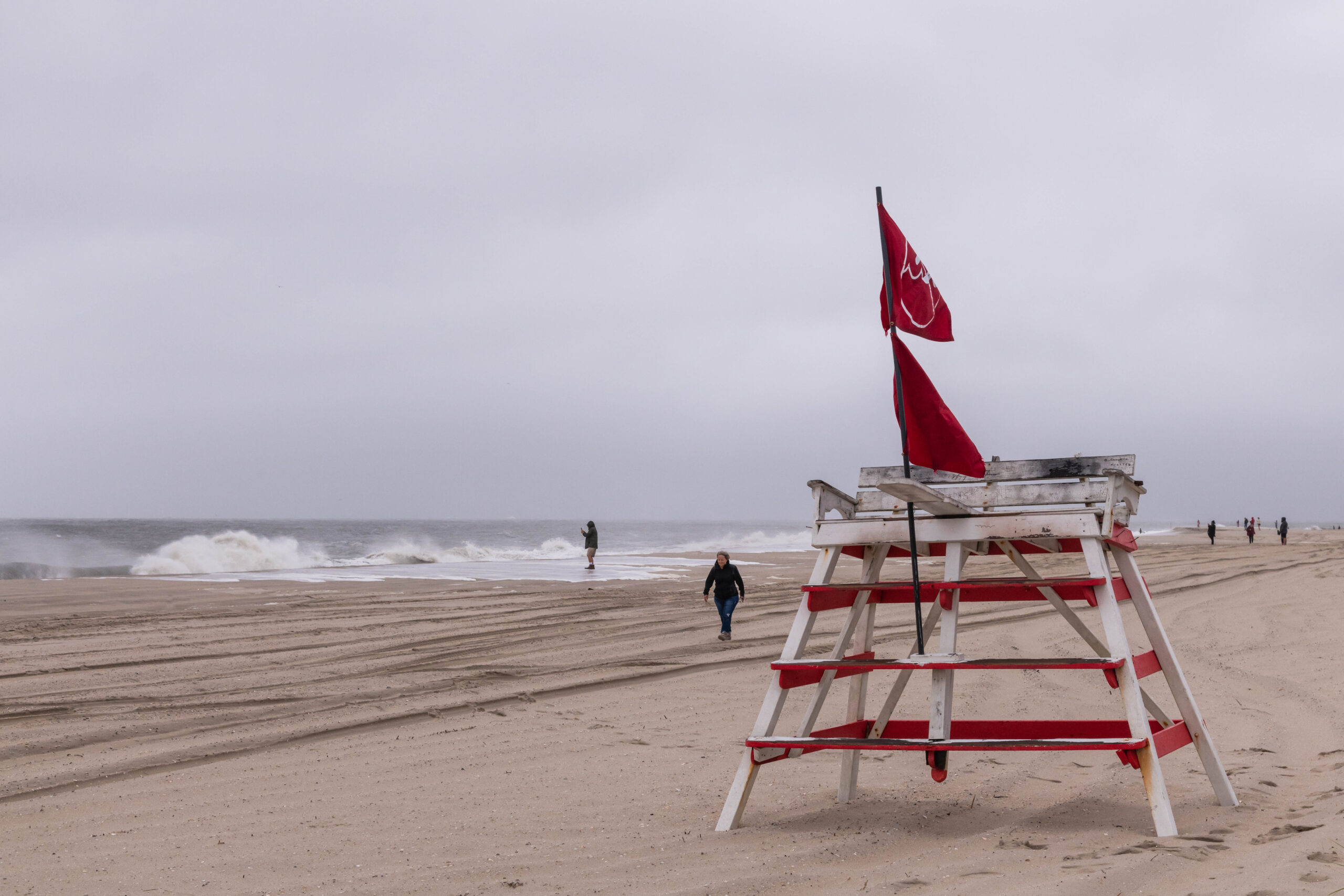 People walking on the beach with the ocean crashing and coming up high on the sand, and a Cape May lifeguard stand with two red "don't swim" flags.