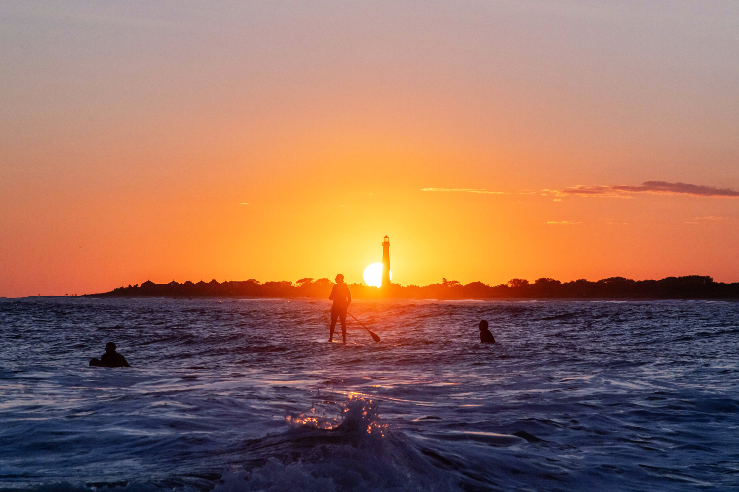 The sun setting directly behind the Cape May Lighthouse with surfers and a paddle boarder in the ocean