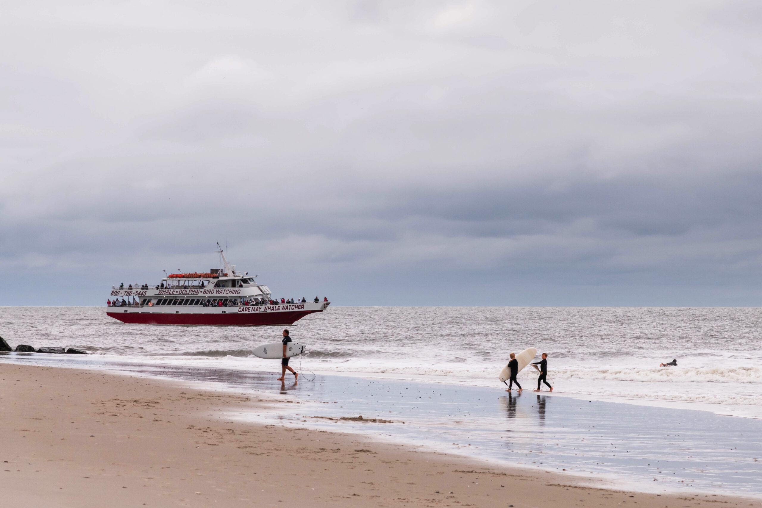 Surfers walking on the beach with the Cape May Whale Watcher boat in the ocean with clouds in the sky