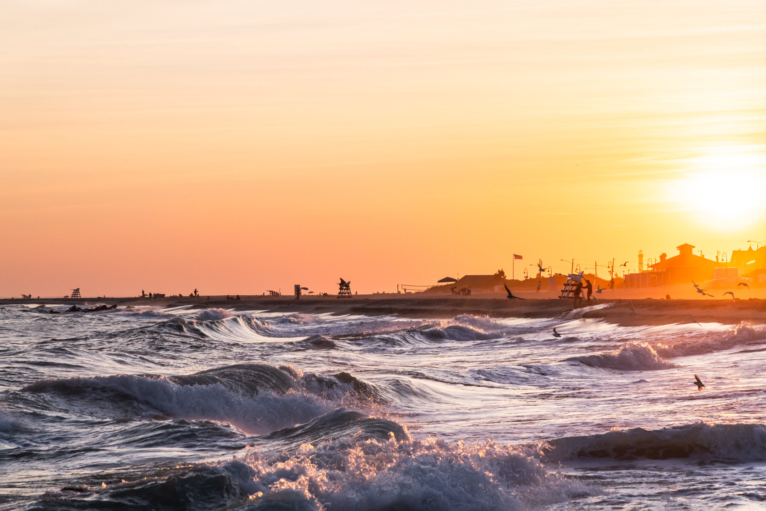 Waves crashing into the beach at sunset with seagulls flying and lifeguard stands dotting the horizon