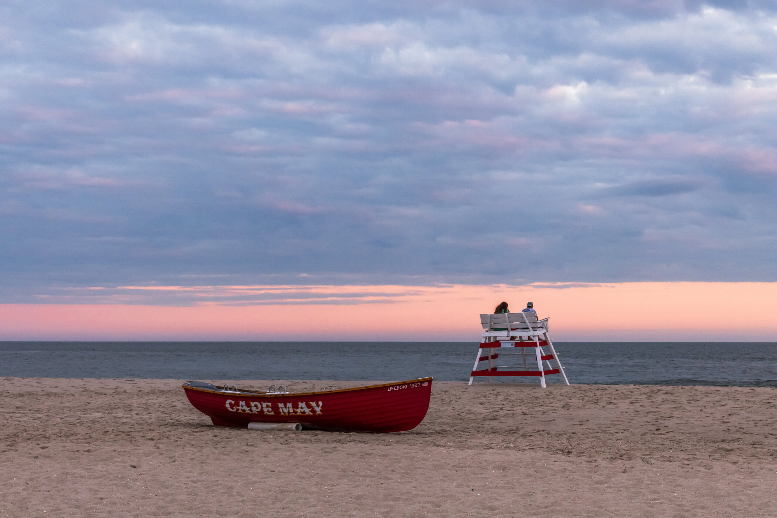 Two people sitting on a lifeguard stand at the beach with a red Cape May lifeguard boat and clouds in the sky with pink at the horizon line