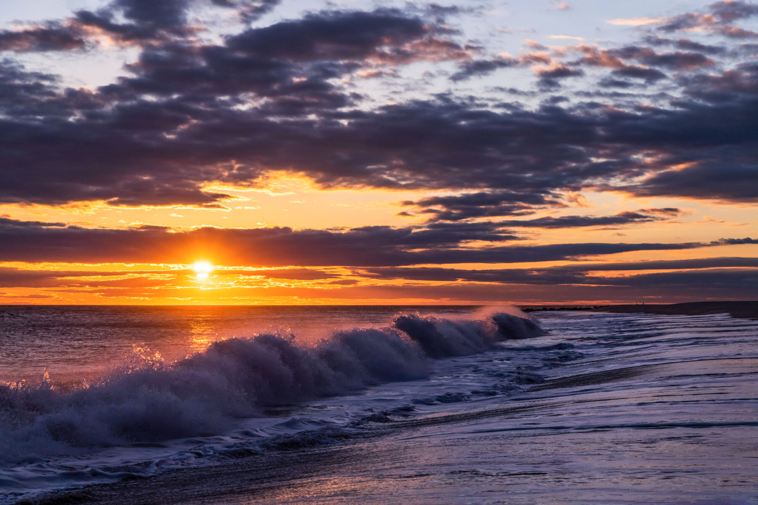 A wave crashing on the beach at sunset with purple, pink, yellow, and orange clouds in the sky