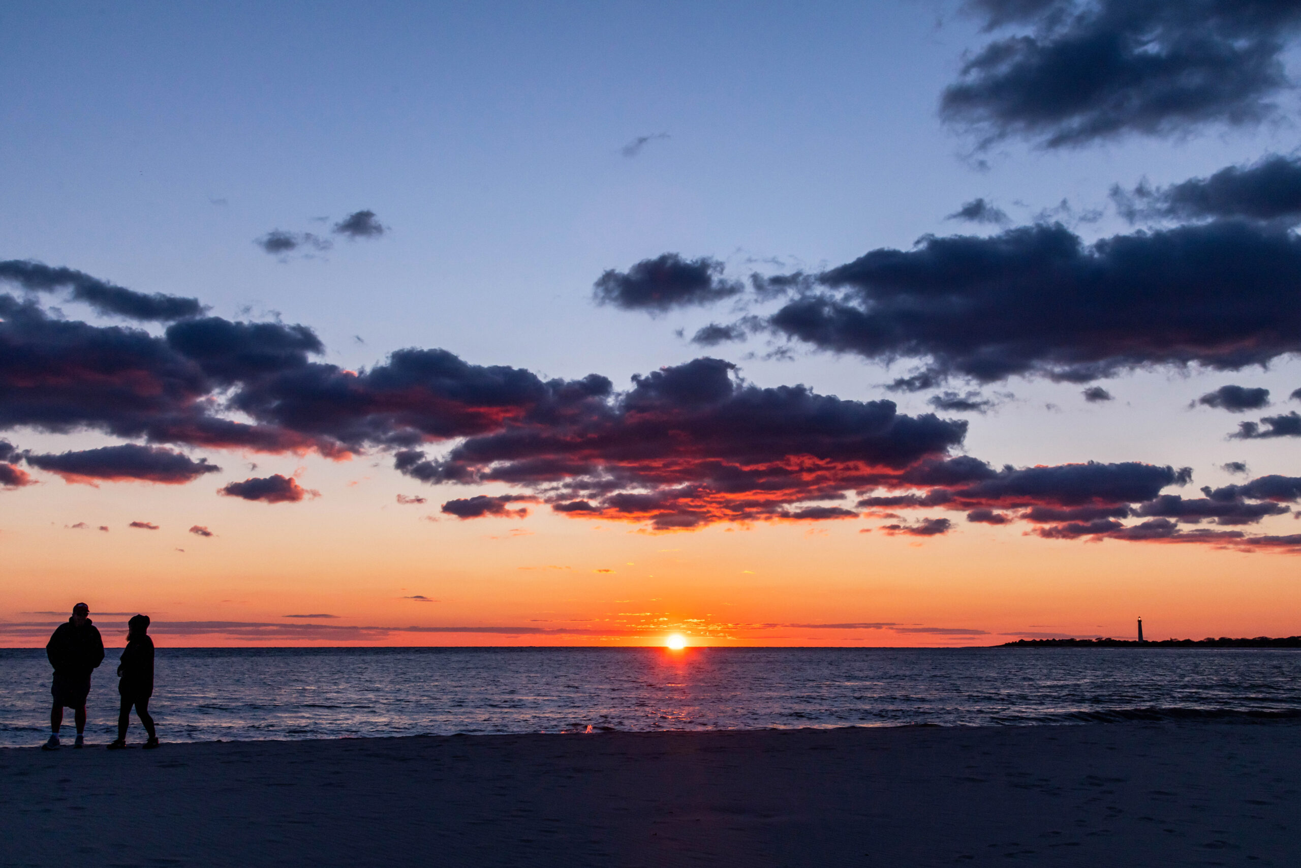 The sun sinking below the horizon with a few purple and pink clouds in the sky, and two people on the beach