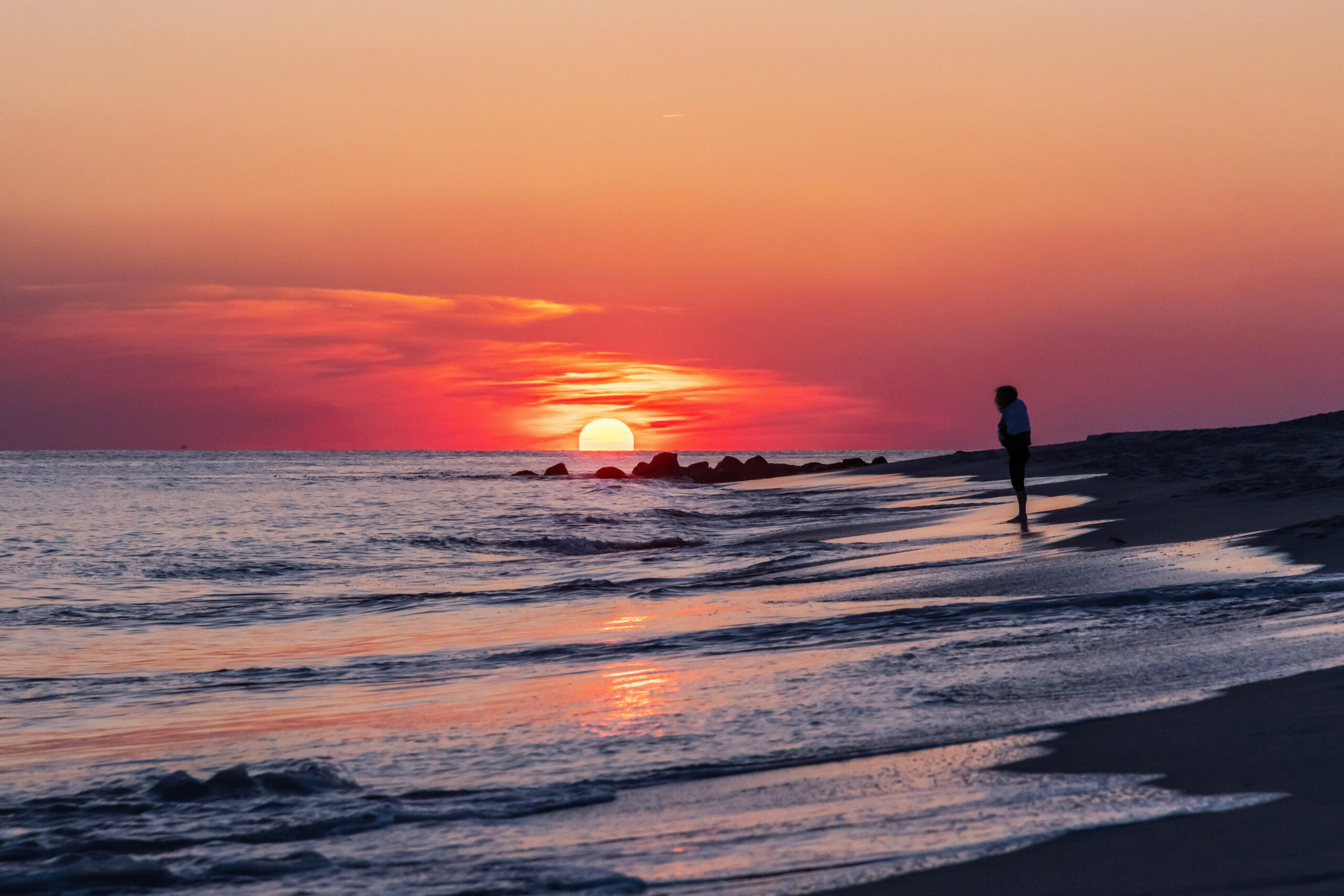 A person standing at the ocean at sunset. The sun and sky look pink and orange, and the tide is rushing in.