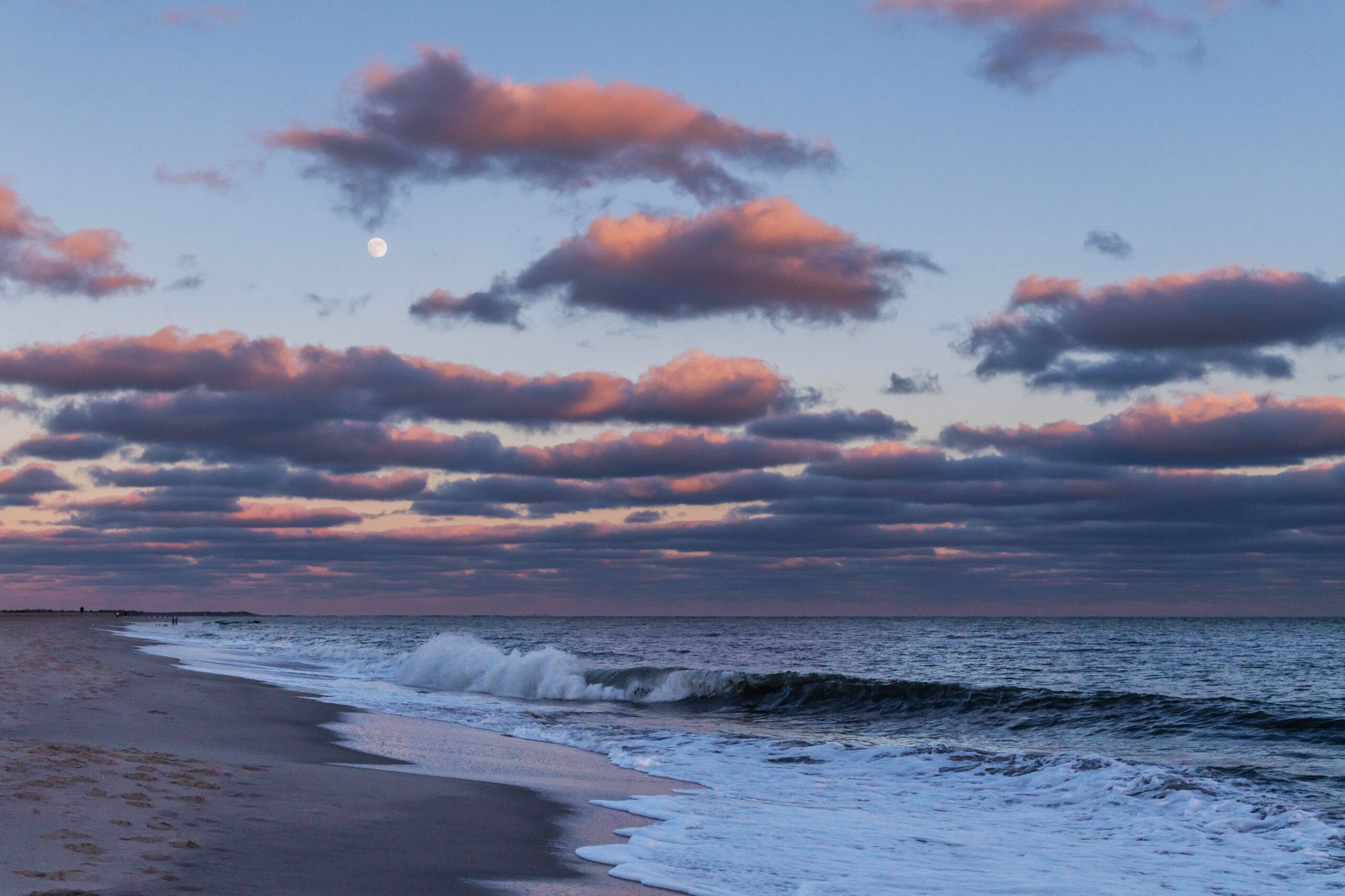 Pink and purple clouds in the sky with a full moon and a wave crashing in the ocean.