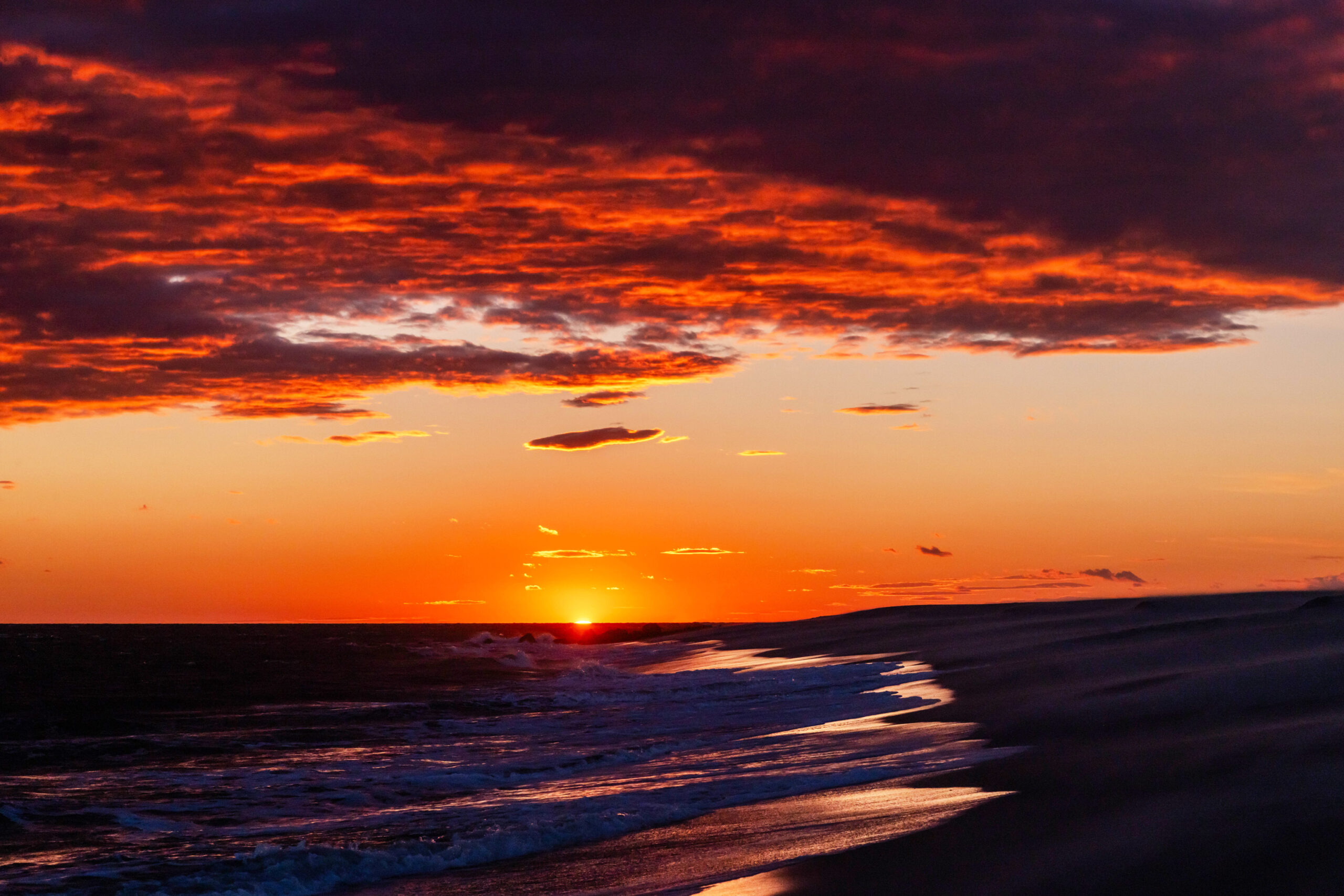 Orange and red clouds in the sky with the sun setting and the light reflected in a dark ocean and the sand