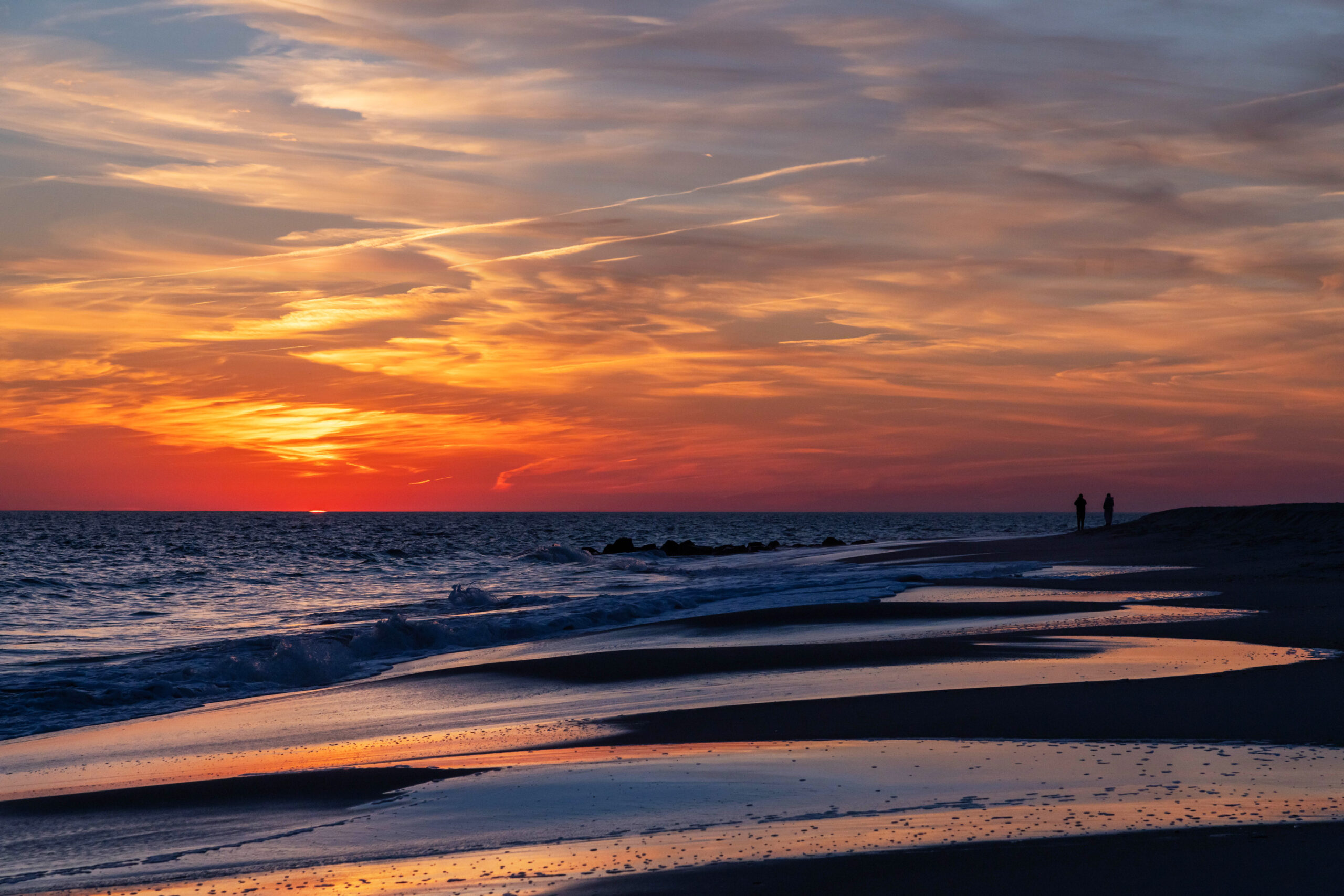 Two people in the distance on the beach watching the sun set behind the horizon. There are thin whispy clouds in the sky. The clouds are orange and red and they are reflected in the ocean and sand.