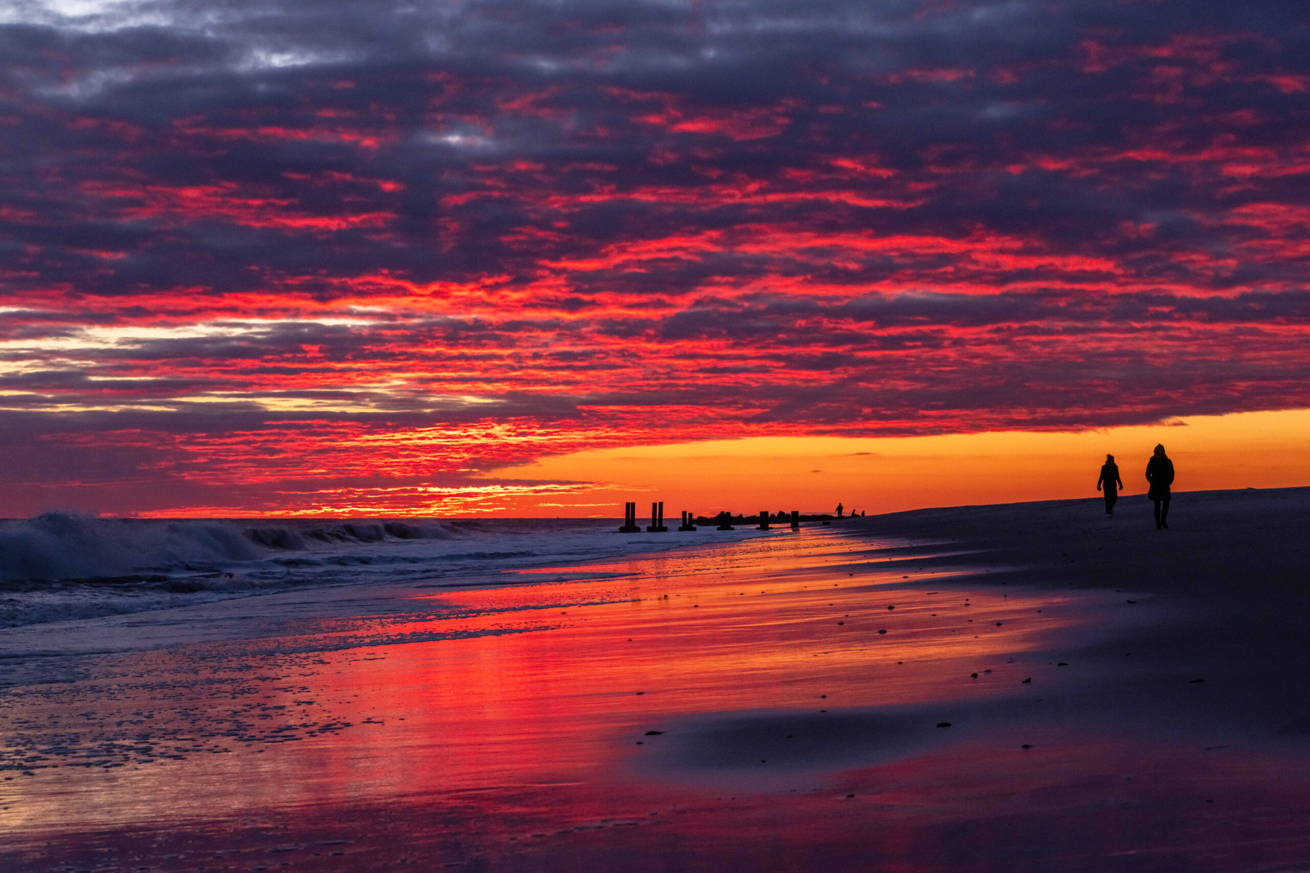 Pink, blue and purple clouds in the sky at sunset and reflected in the ocean and shoreline. Two people are walking next to the ocean.