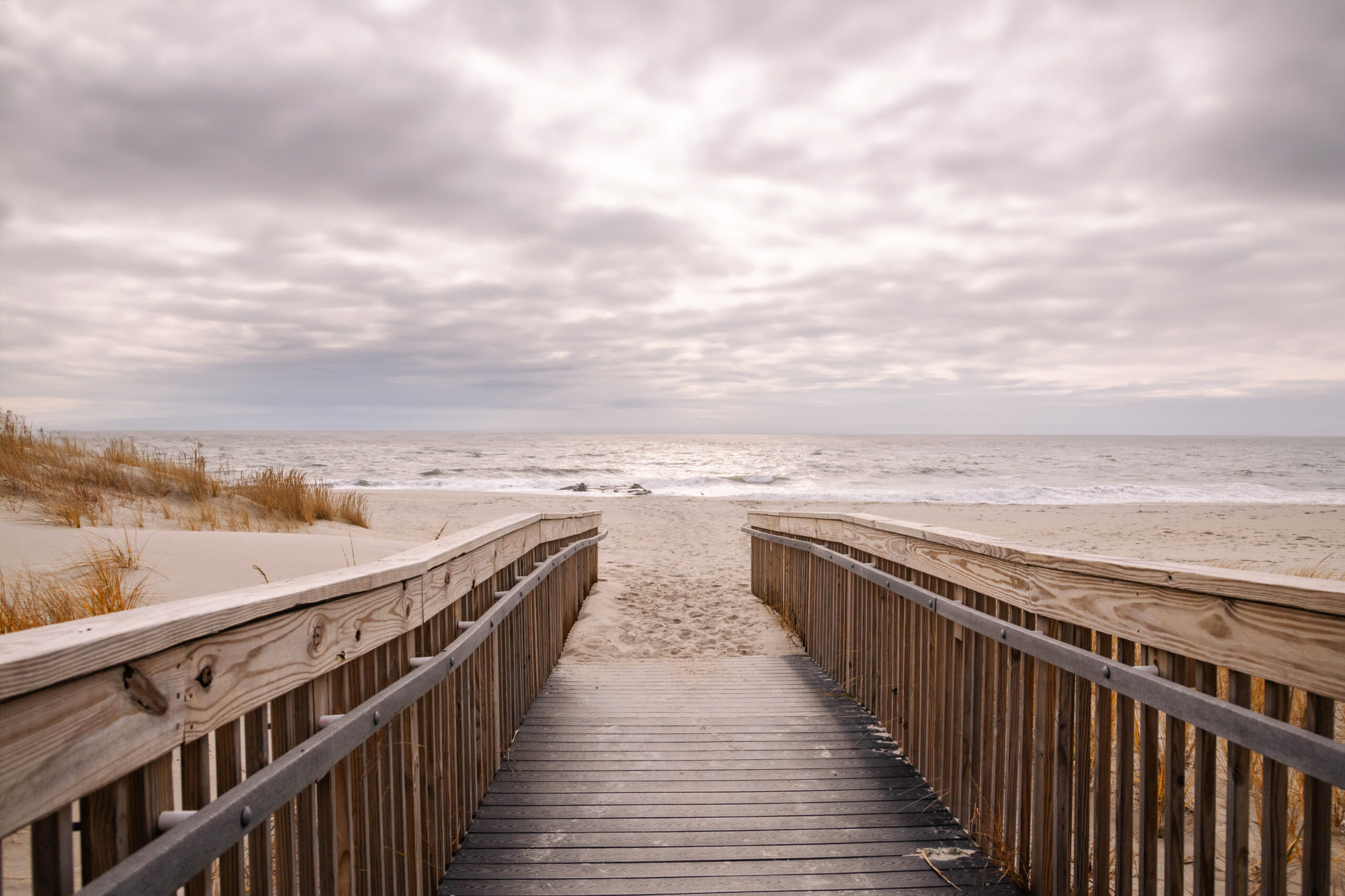 A wooden path leading to the beach with a cloudy sky and hints of sunlight reflected on the ocean
