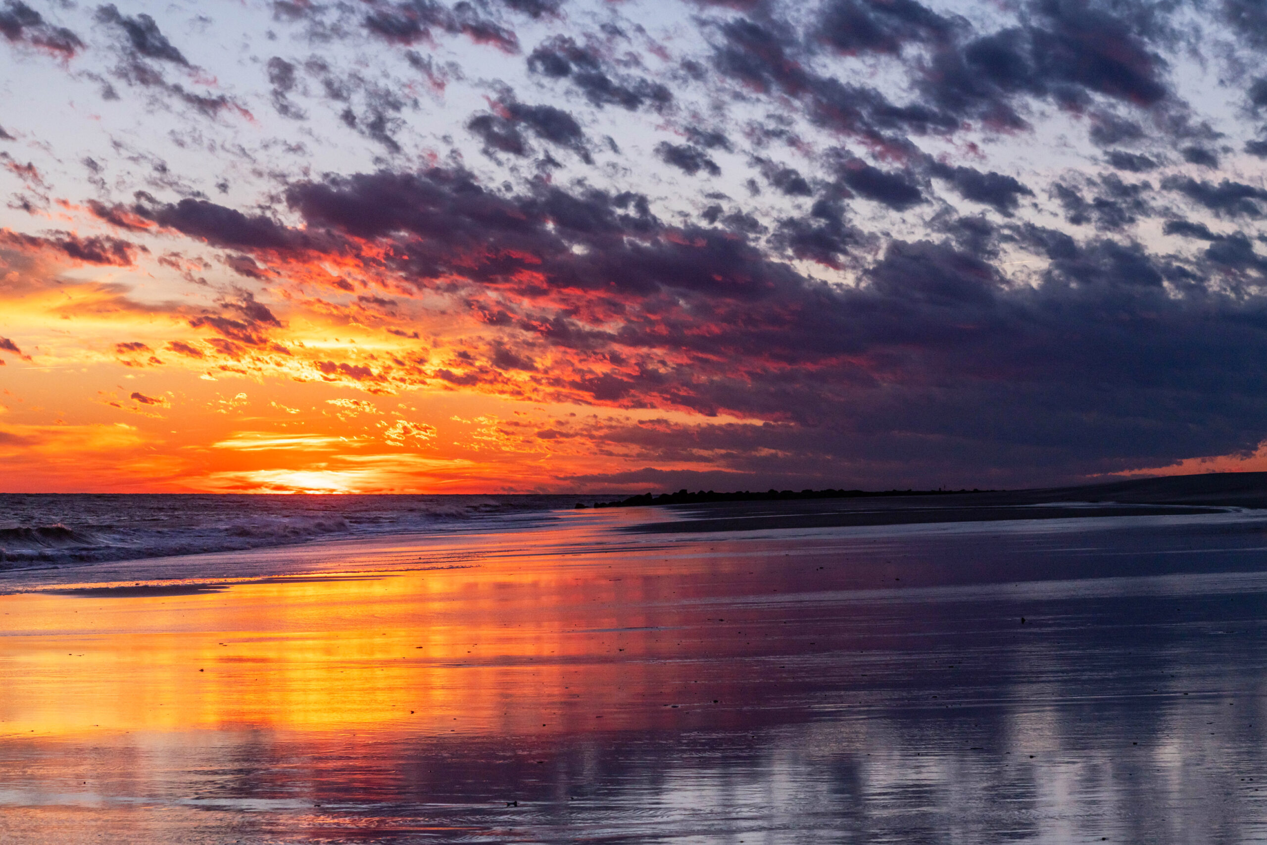 Purple, pink, and orange clouds in the sky at sunset. The clouds are reflected in the shoreline and ocean.