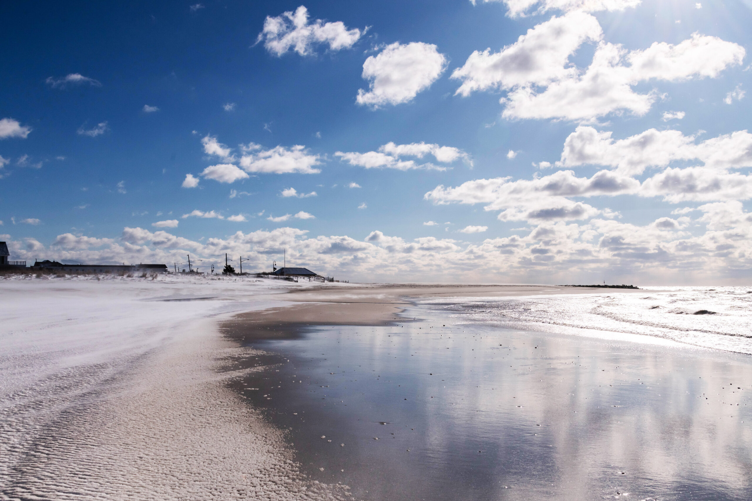 Snow on the beach with puffy clouds in a blue sky. The sky and clouds are reflected in the ocean and shoreline.