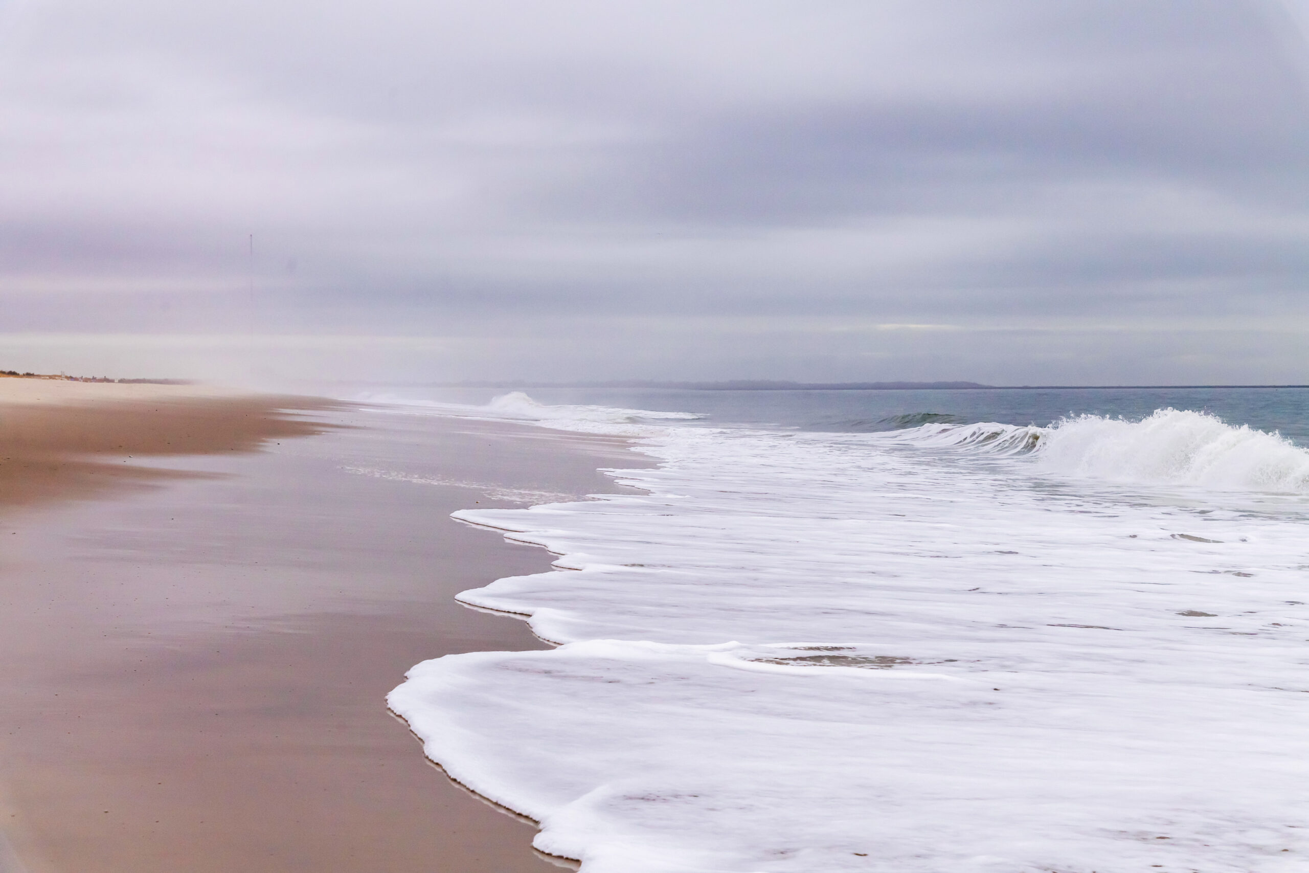 The ocean rushing into the beach with a cloudy sky and fog in the distance