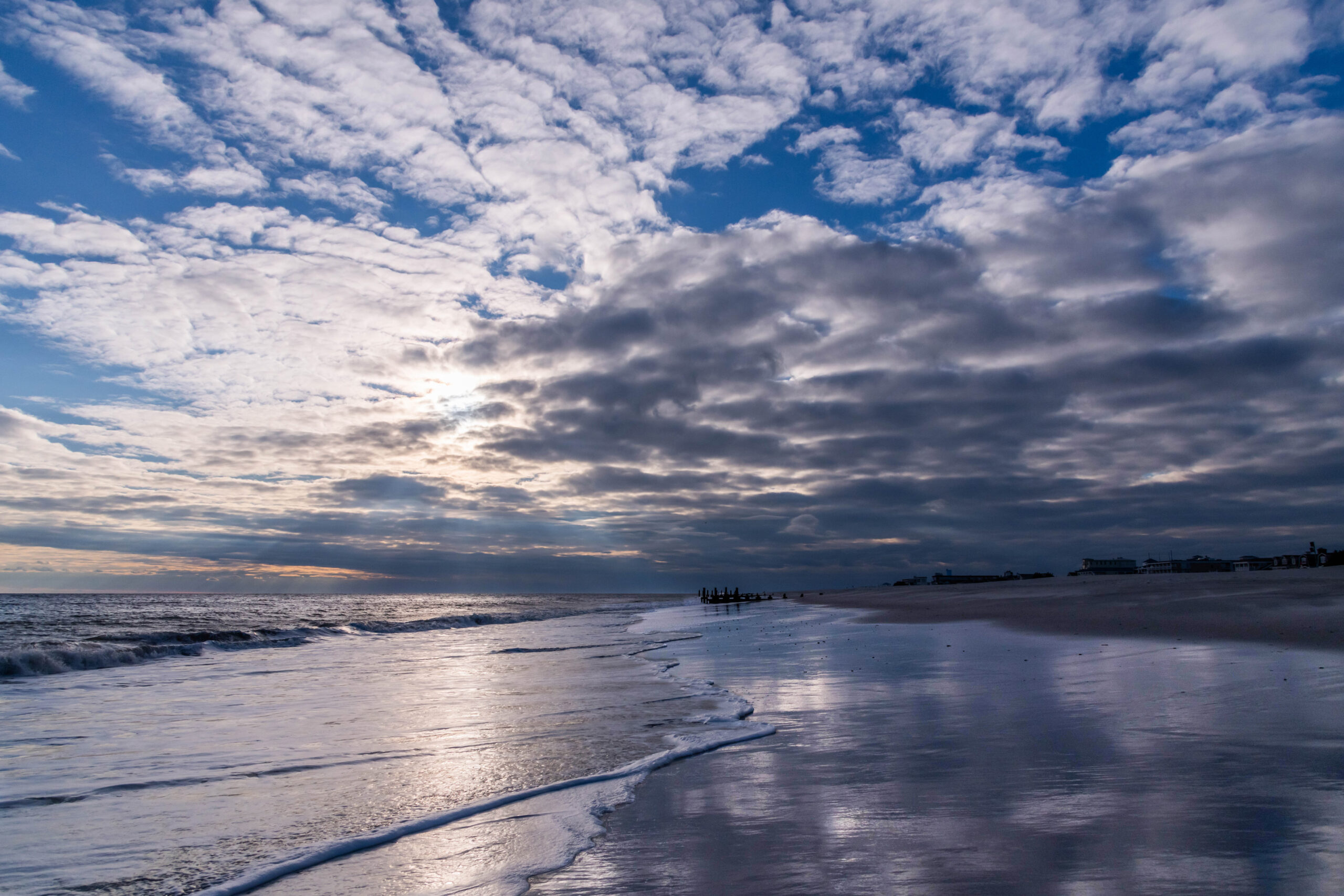 The ocean rushing into the beach with the sun and blue sky breaking through clouds. The sky and clouds are reflected in the shoreline.