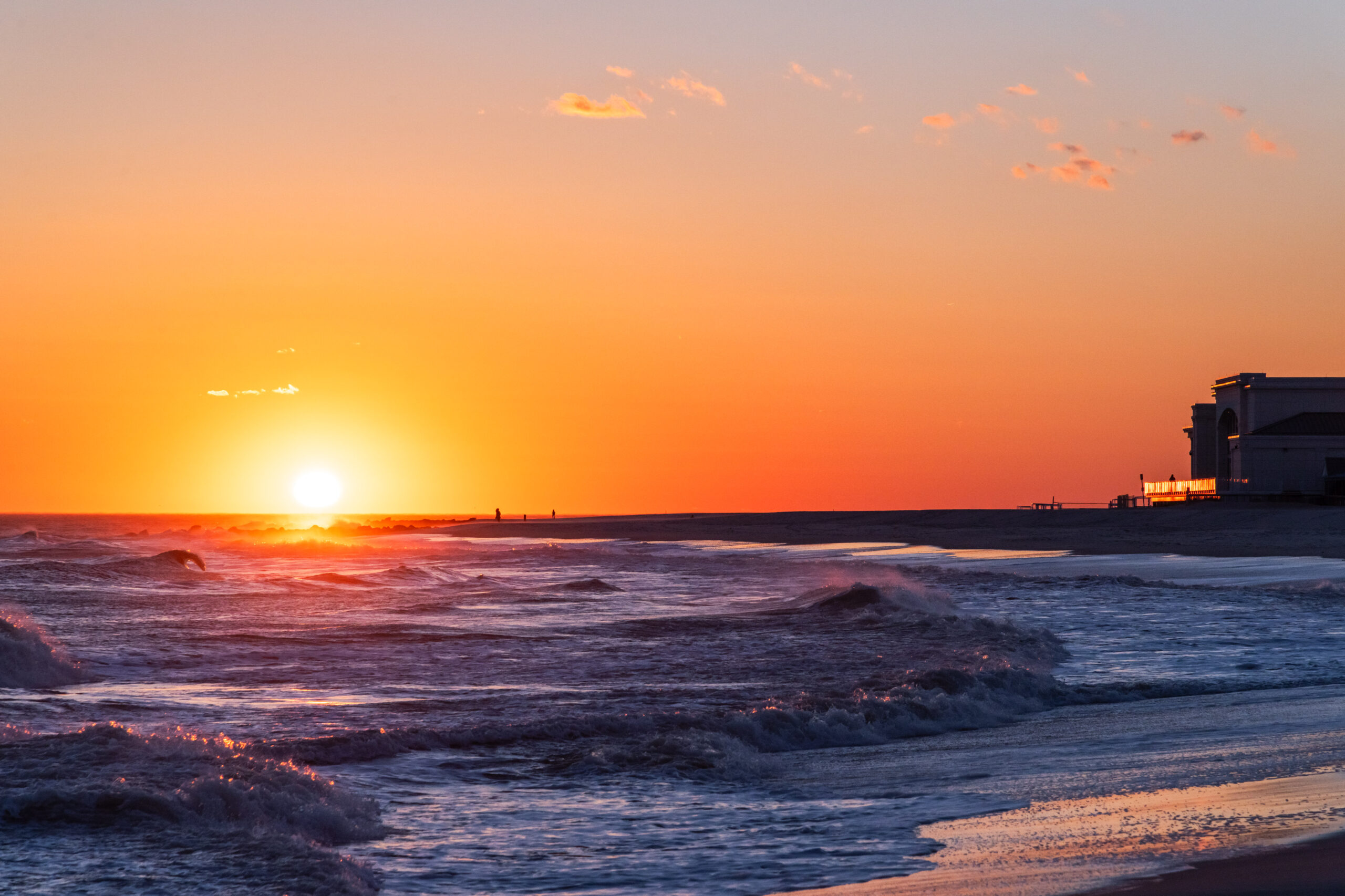 The sun setting in a clear orange sky with waves crashing on the beach. The sun is shining off of the Cape May Convention Center in the distance.