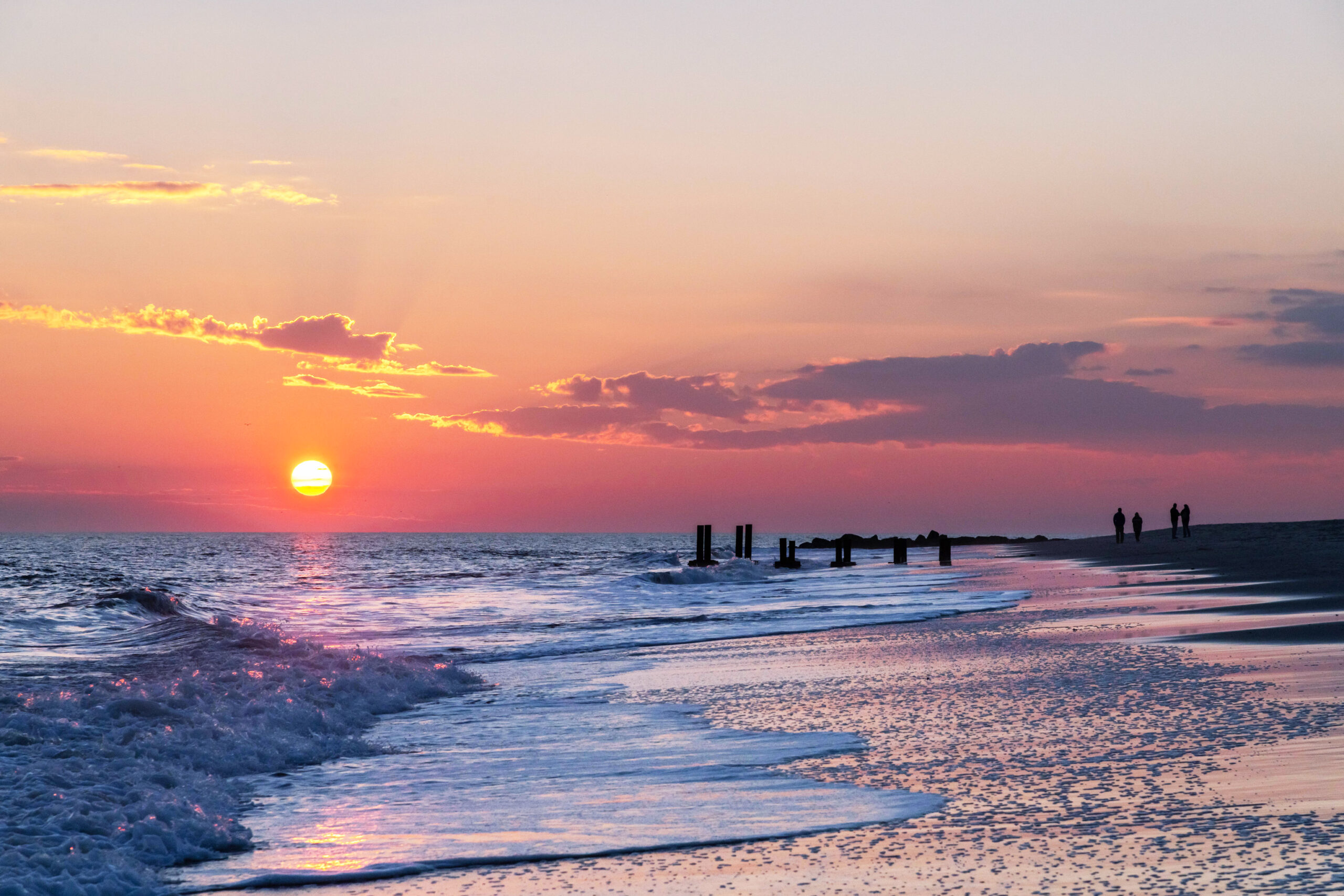 The sun setting with waves rolling into the shoreline and couples walking on the beach. There are a few clouds in the sky.