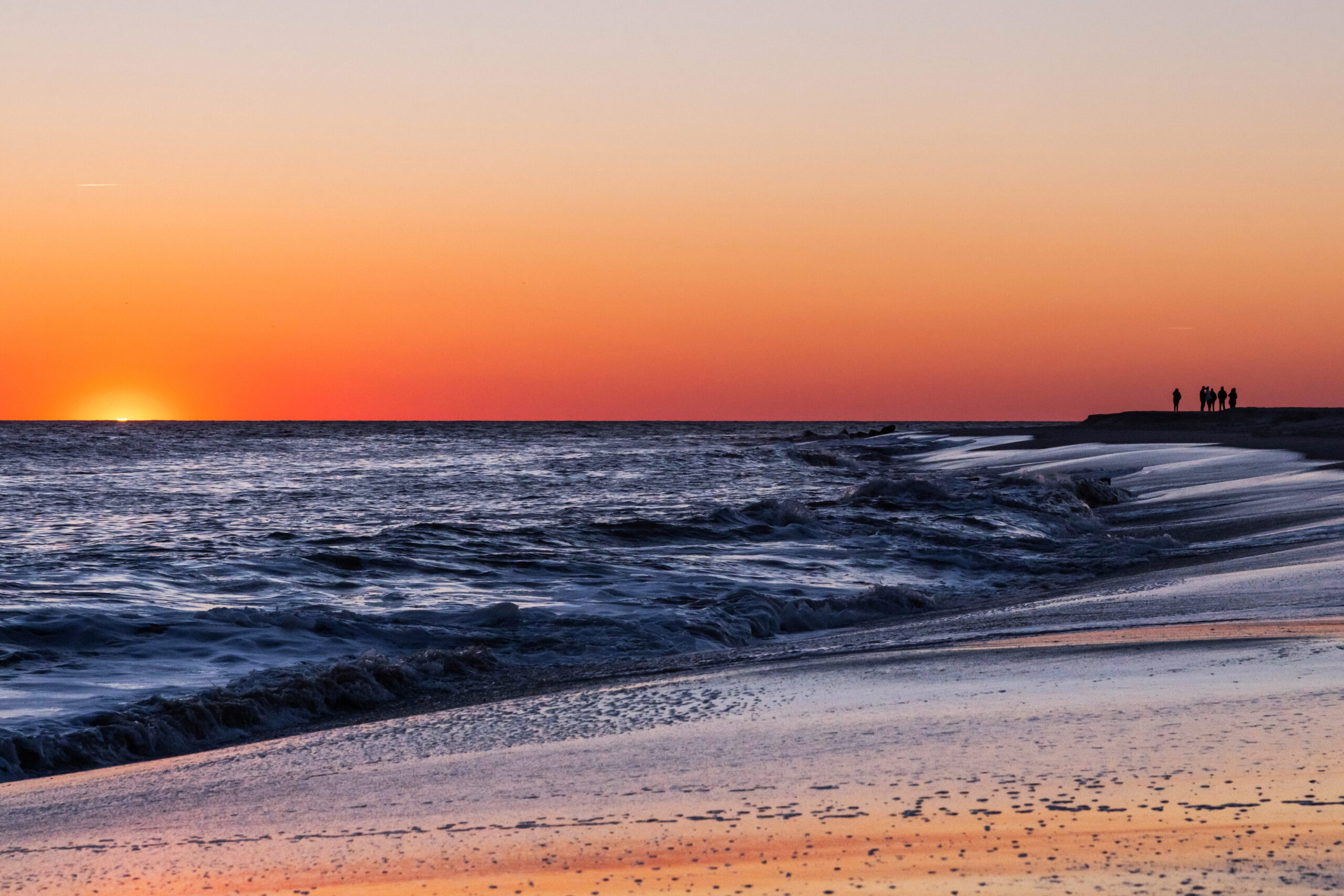 A group of people in the distance standing by the ocean with the sun setting. There is a clear orange sky, and the sky is reflected in the sand and shoreline.