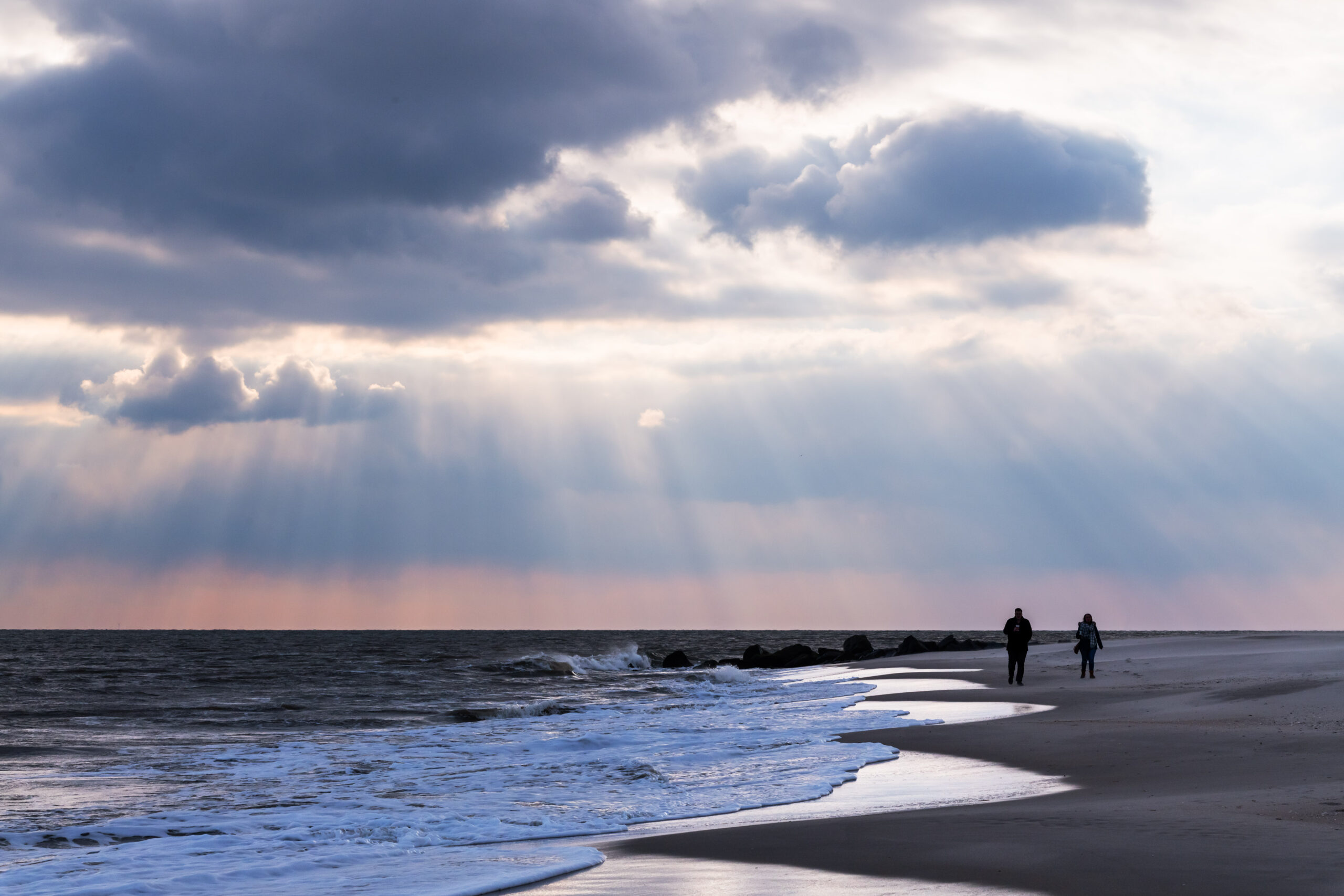 Sun streaming through big dark clouds in the sky. There are two people walking along the ocean at the beach.