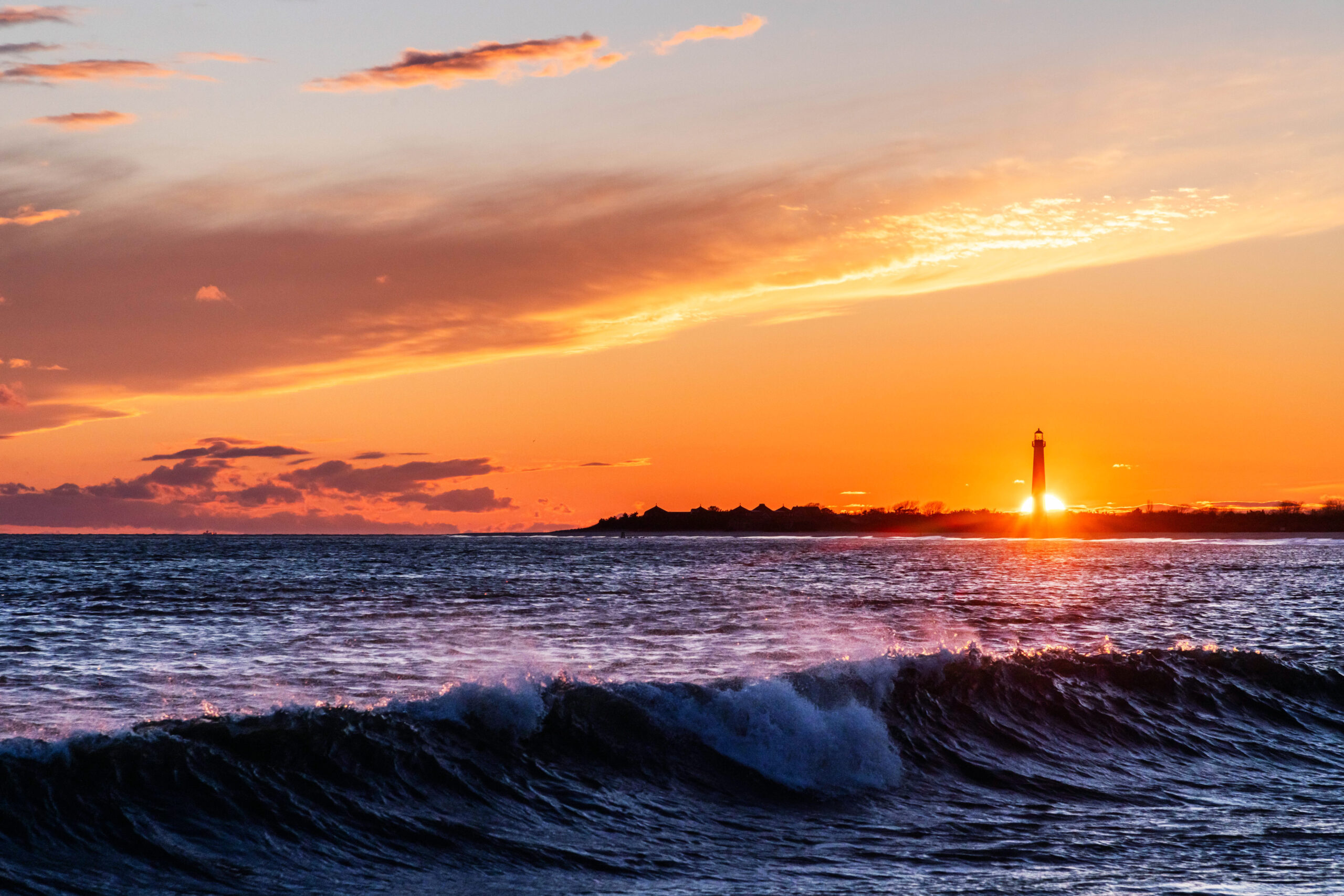 The sun setting directly behind the lighthouse with an orange sky and a wave crashing in the ocean