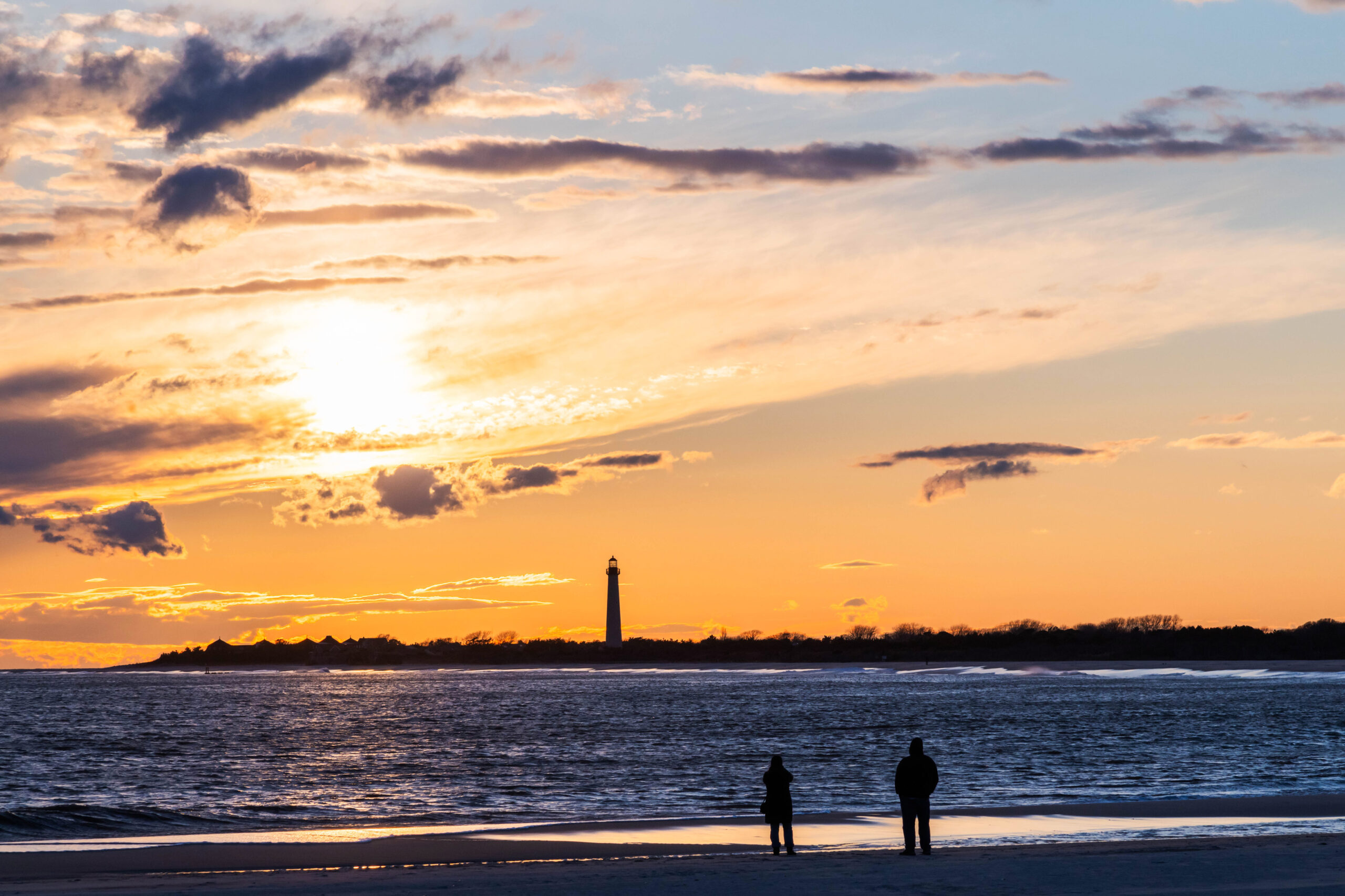 Two people watching the sun set on the beach. The sun is behind thin wispy clouds, and the Cape May Lighthouse in the distance. The sky is orange and blue.