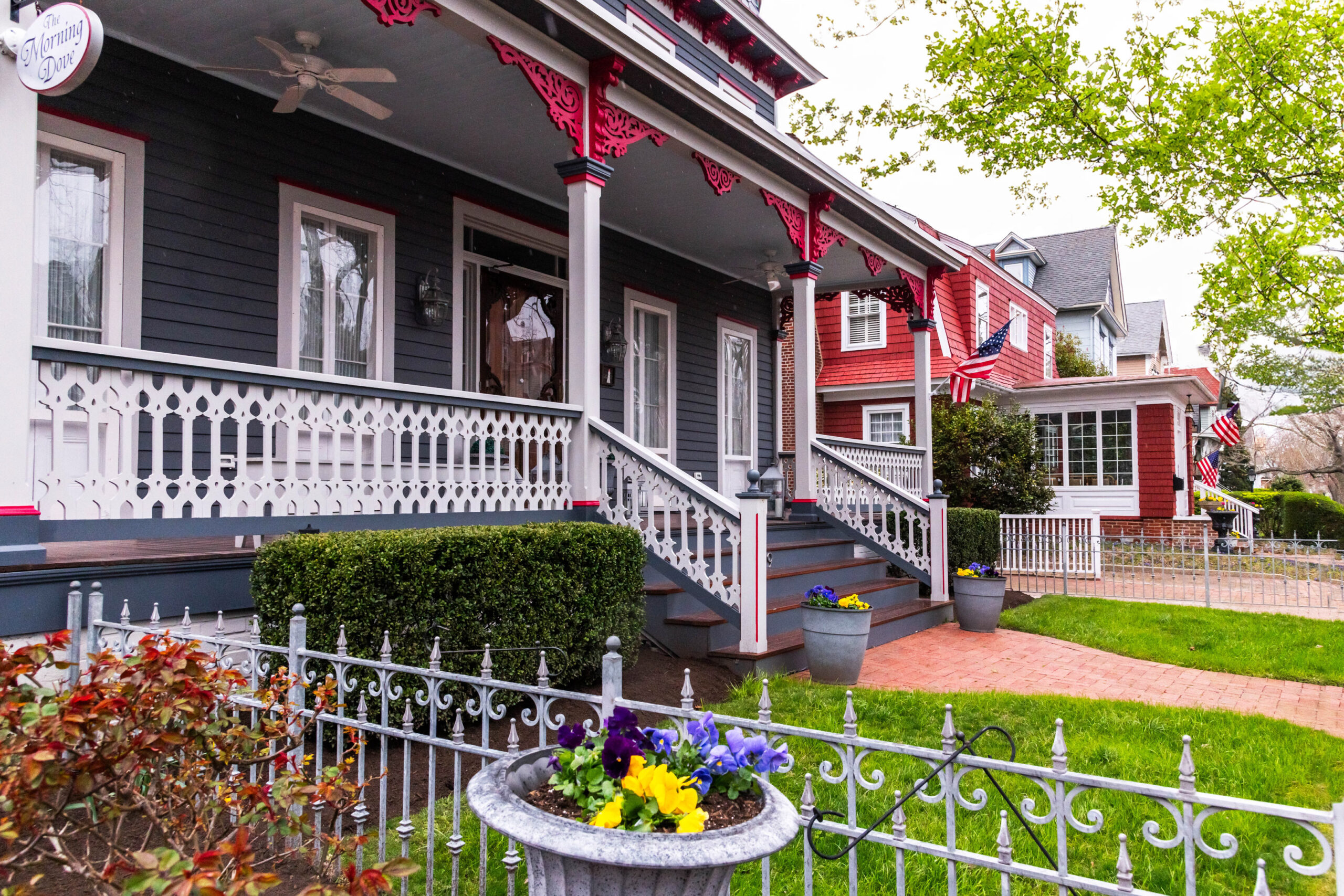 A grey blue Victorian porch with flower pots filled with yellow and purple pansies. It's a cloudy day, and the other houses on Hughes street are in the background.