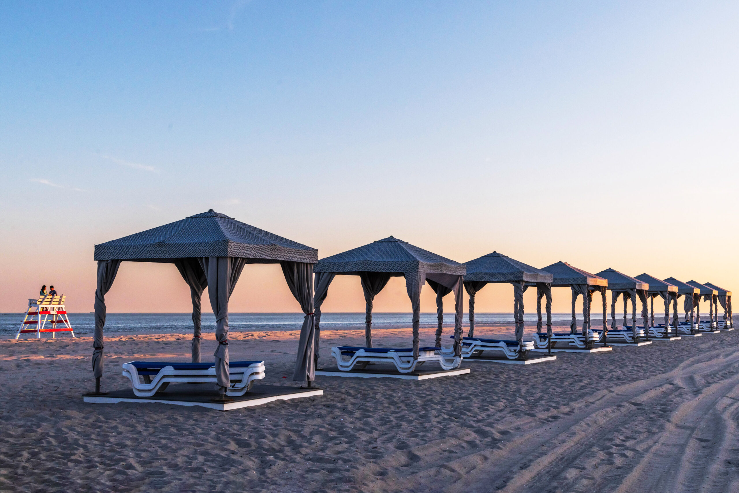 A row of empty beach cabanas at sunset on the beach. There are two people sitting on a Cape May lifeguard stand in the background.