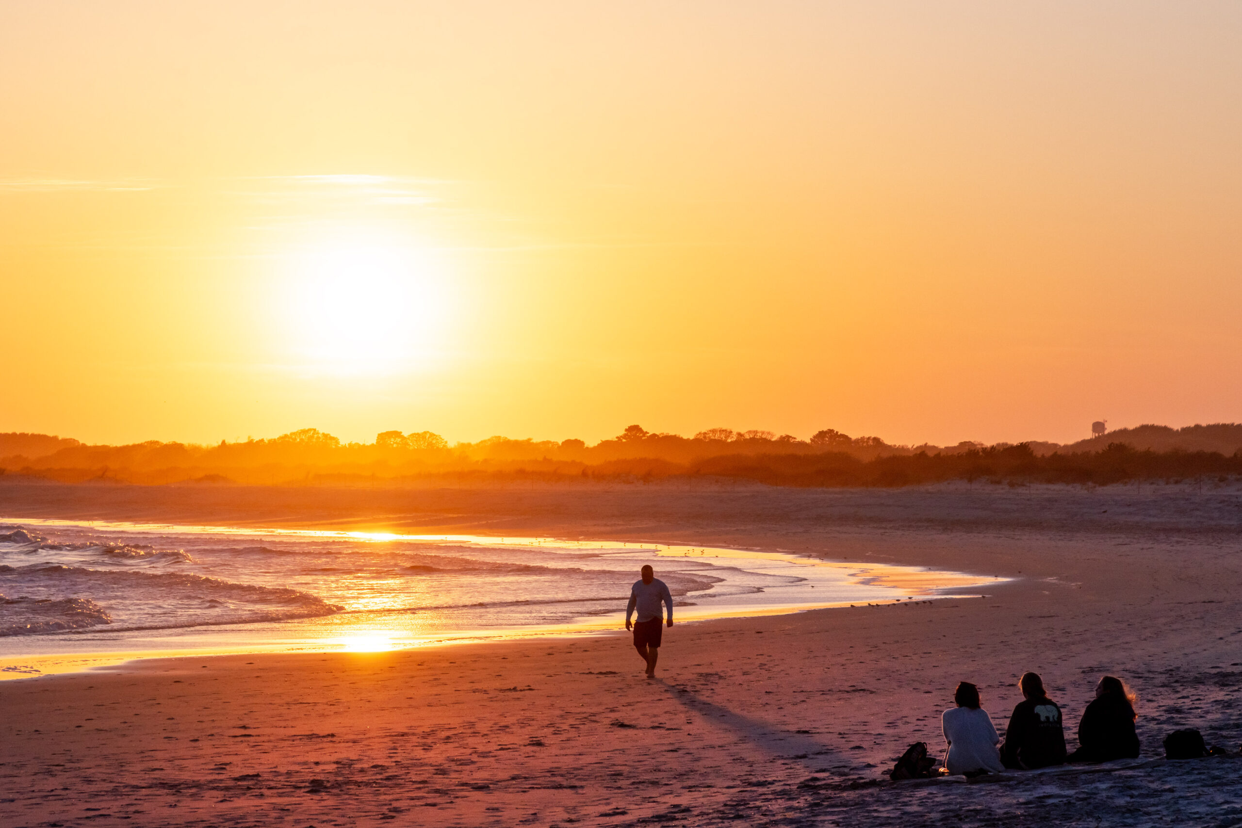A wide view of sunset at the beach. The sky is golden orange. A person is walking by the water, and three people are sitting together on the sand looking at the sun.
