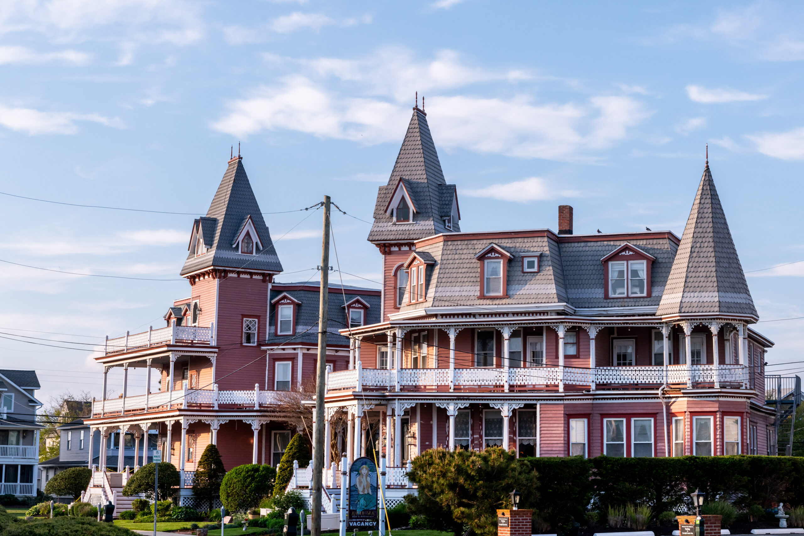 A wide view of the Angel of the Sea, two Victorian houses with pink siding, white porch railings, and gray roofs. There is a blue sky with a few small wispy white clouds.