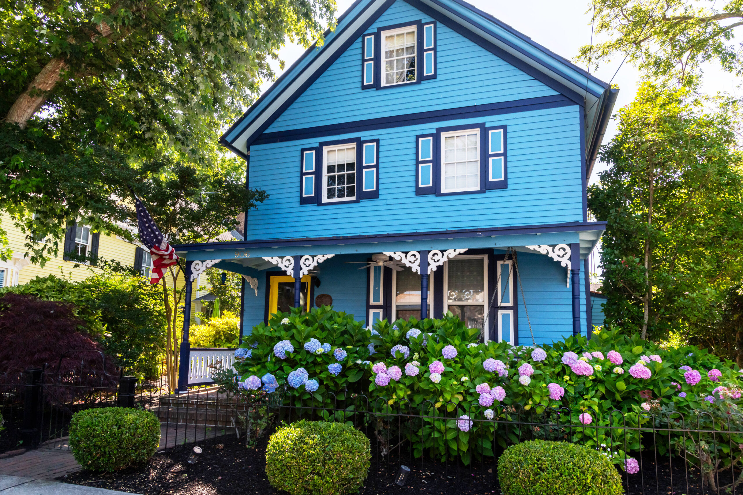 A wide view of a bright teal Victorian style home with bushes of hydrangea in front of the front porch. 