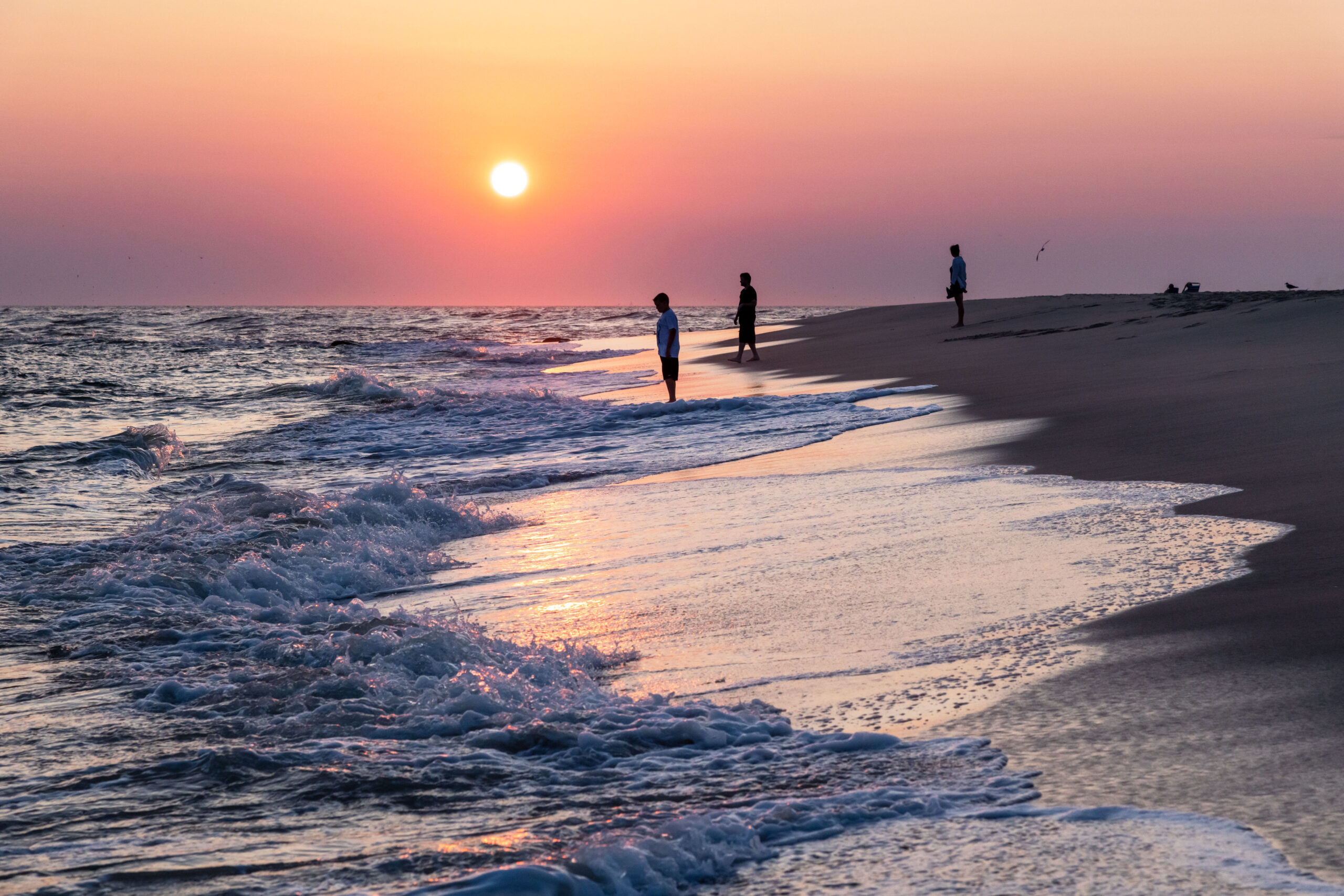 A wide view of the shoreline at the beach at sunset. There is a wave rushing into the beach, and three people are in silhouette standing by the water. There is a clear sky with pink, purple, and yellow.