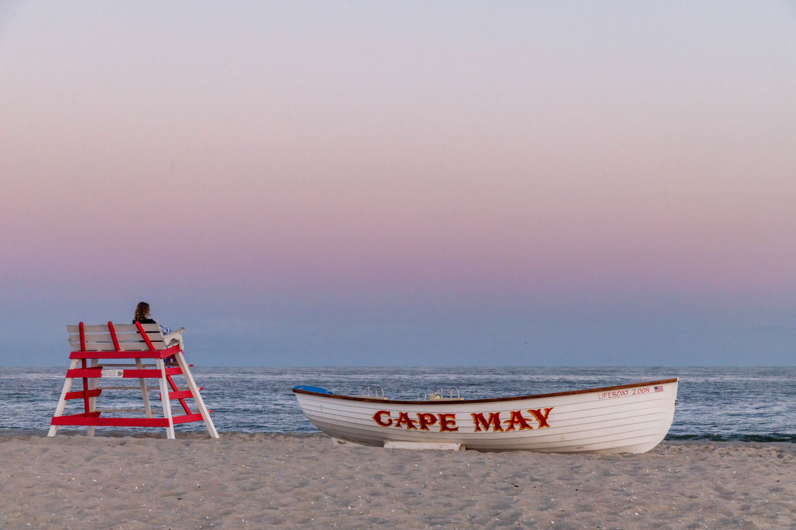 A wide view of a person sitting on a red and white lifeguard stand and a Cape May lifeguard boat on the beach at sunset. The sky is purple, pink, and light blue. The boat is white with "CAPE MAY" written in red.