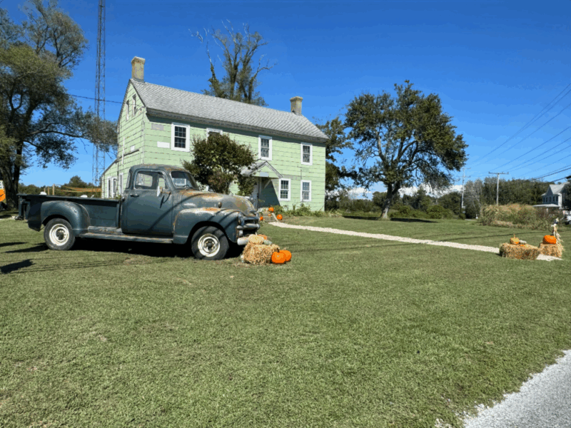 Blue skies behind an old farm house with a vintage truck, hay bales and pumpkins decorating the yard