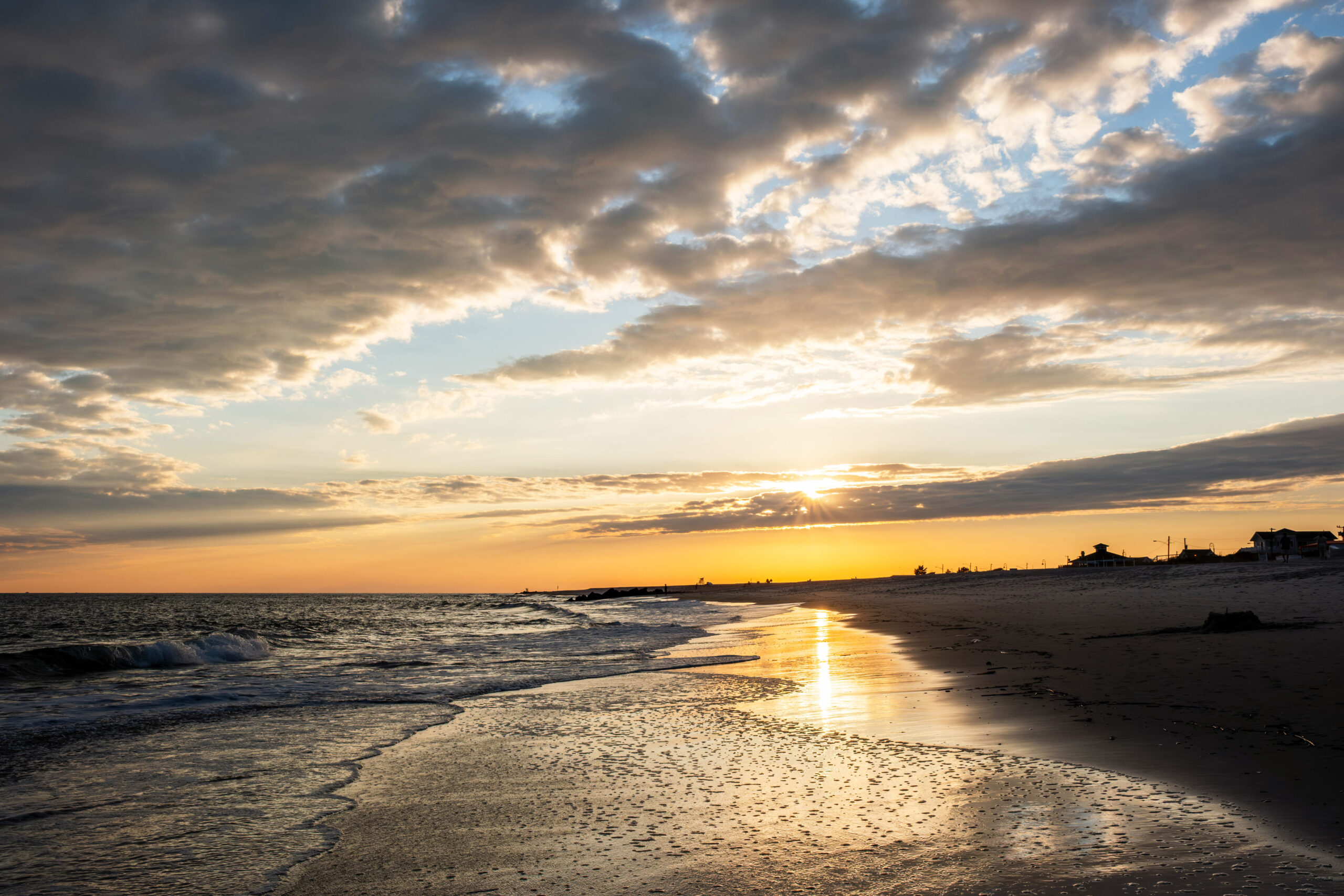 A wide view of sunset at the beach. There are thin clouds in the sky, and the sun is shinning behind one. Blue and orange sky is breaking up the clouds. The sunlight is reflected in the ocean and shoreline.