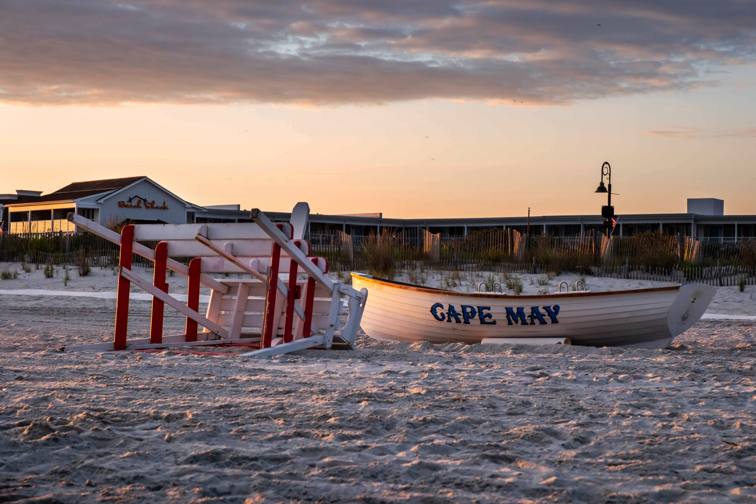 A wide view of a lifeguard stand and a Cape May lifeguard boat on the beach at sunset. The lifeguard stand is down on its side. There is a hotel and beach fence pictured behind the boat and lifeguard stand. "CAPE MAY" is written on the boat.