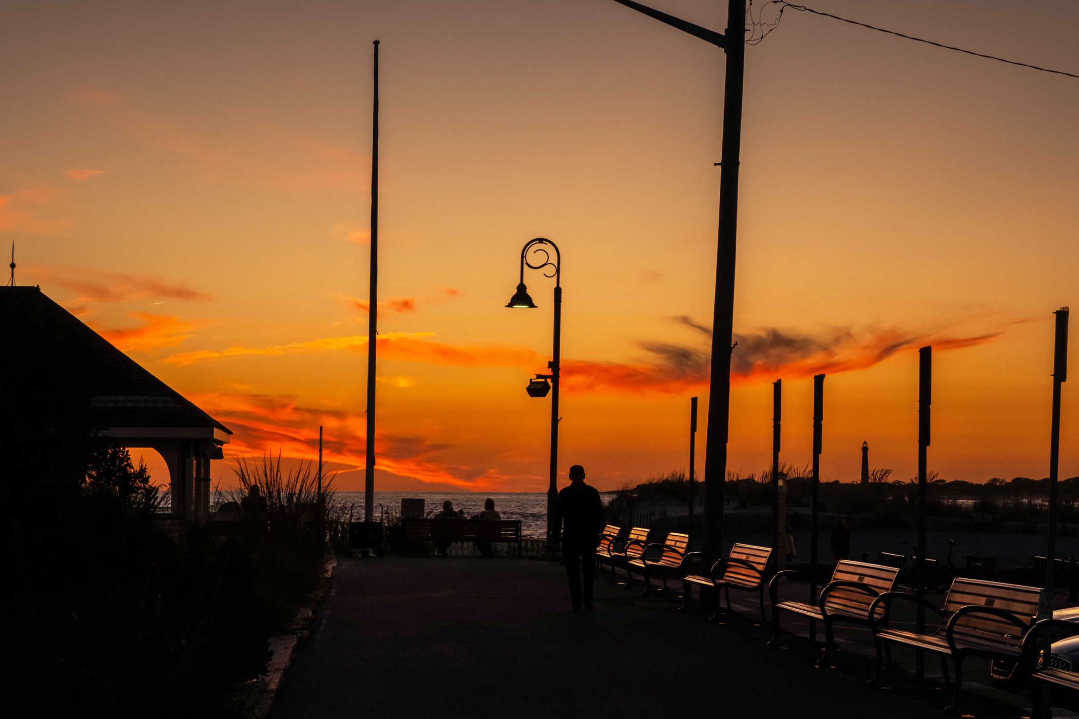 A wide view of a person walking on the promenade at sunset. The person and the promenade are mostly in silhouette, with the benches on the promenade reflecting orange light from the sky. The sky is bright orange and yellow.