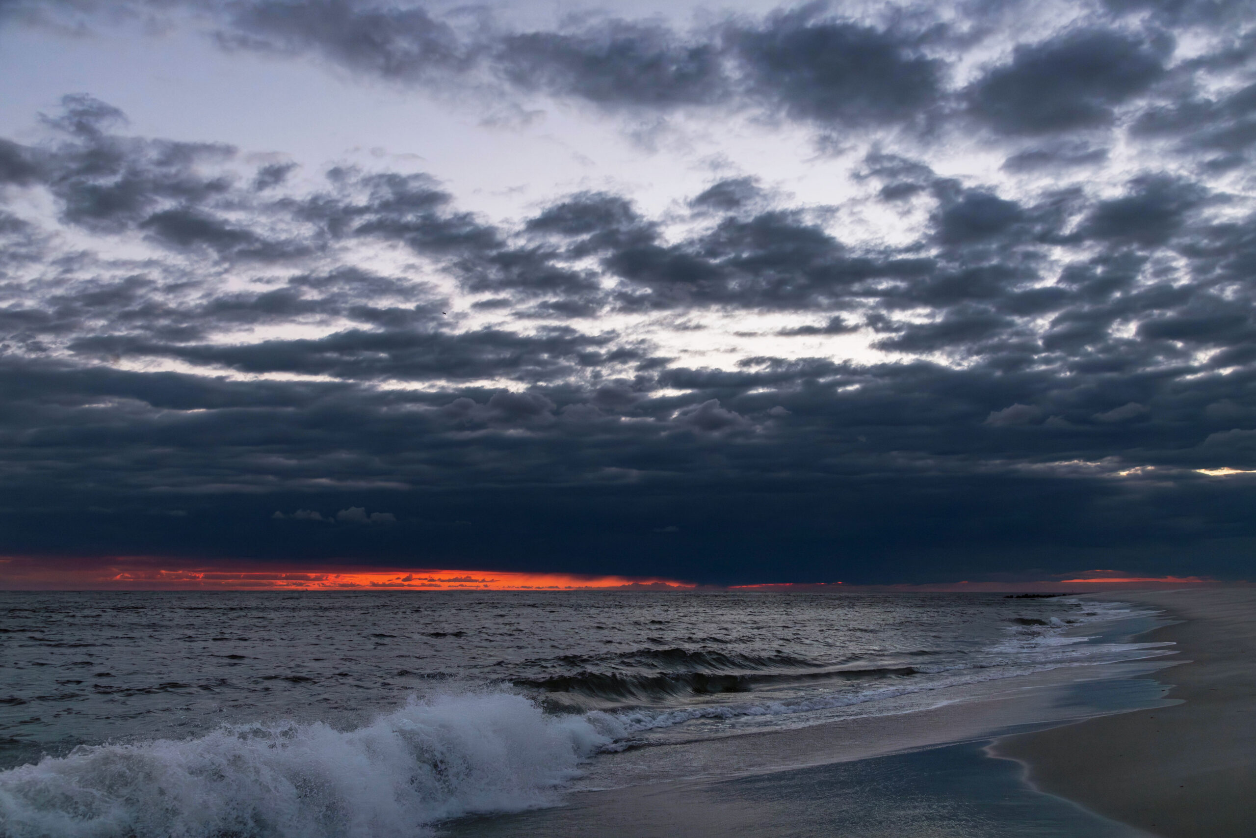 A wide view of sunset at the beach with a wave crashing. There are dark purple thin clouds in the sky. Some sky is breaking up the clouds, and there is a bright thin stripe of orange light from sunset at the horizon.