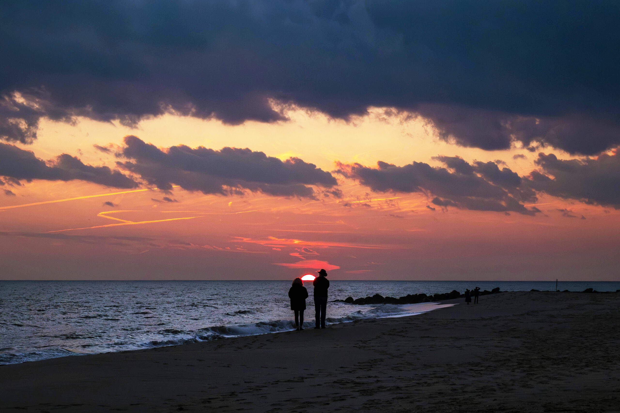 A wide view of two people standing on the beach watching sunset. The sun is setting in between and behind the two people. There are a few dark purple puffy clouds in the sky. The horizon and sun are pink.