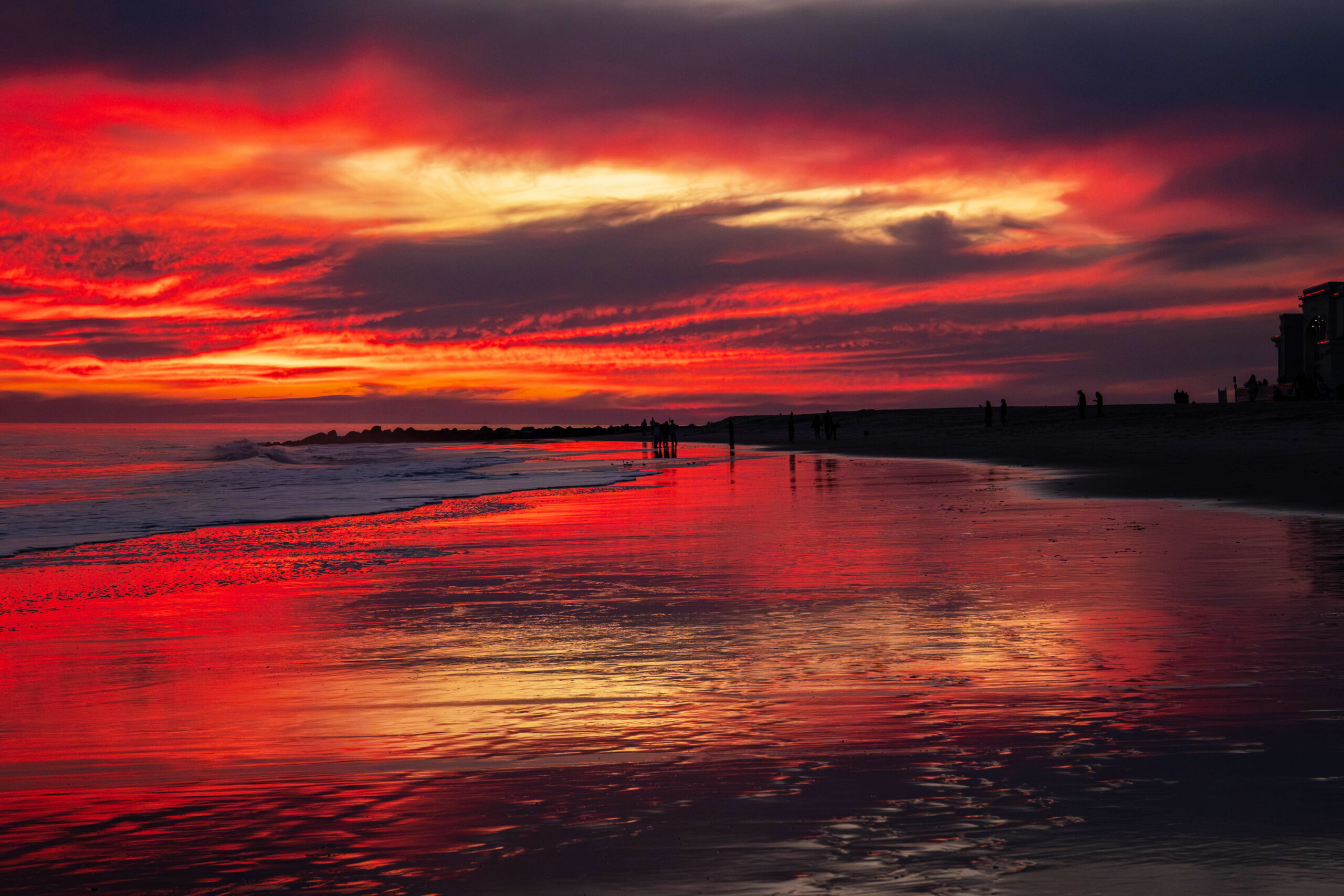 A wide view of a brilliantly colorful sunset at the beach. There are dark purple, red, and orange clouds int he sky. The colors are reflected in the ocean and shoreline. 
