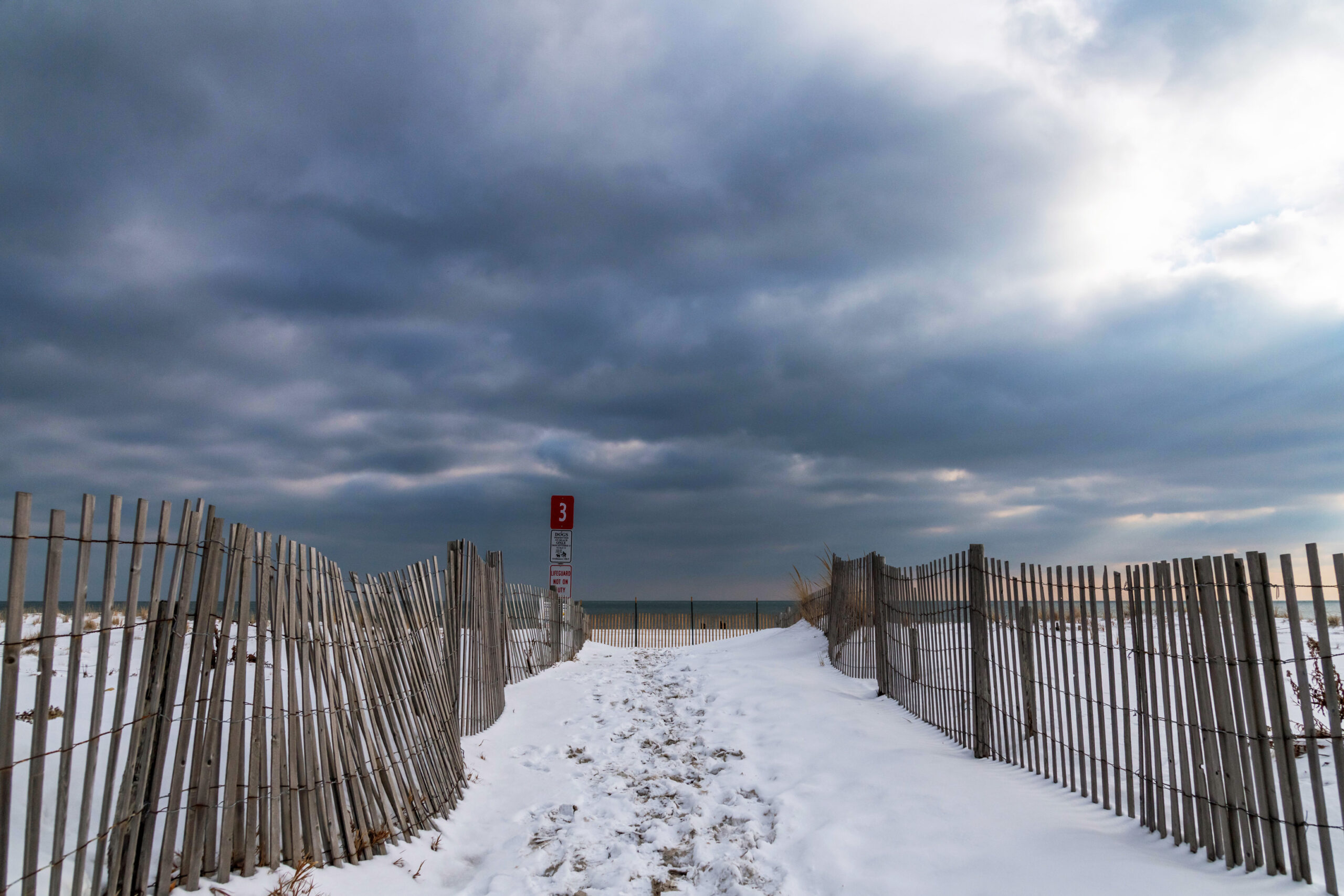 A wide view of an entrance to the beach with snow on the sand. The entrance is marked by a brown fence on either side. There is a red sign with the number three at the entrance. There are dark blue puffy clouds in the sky.