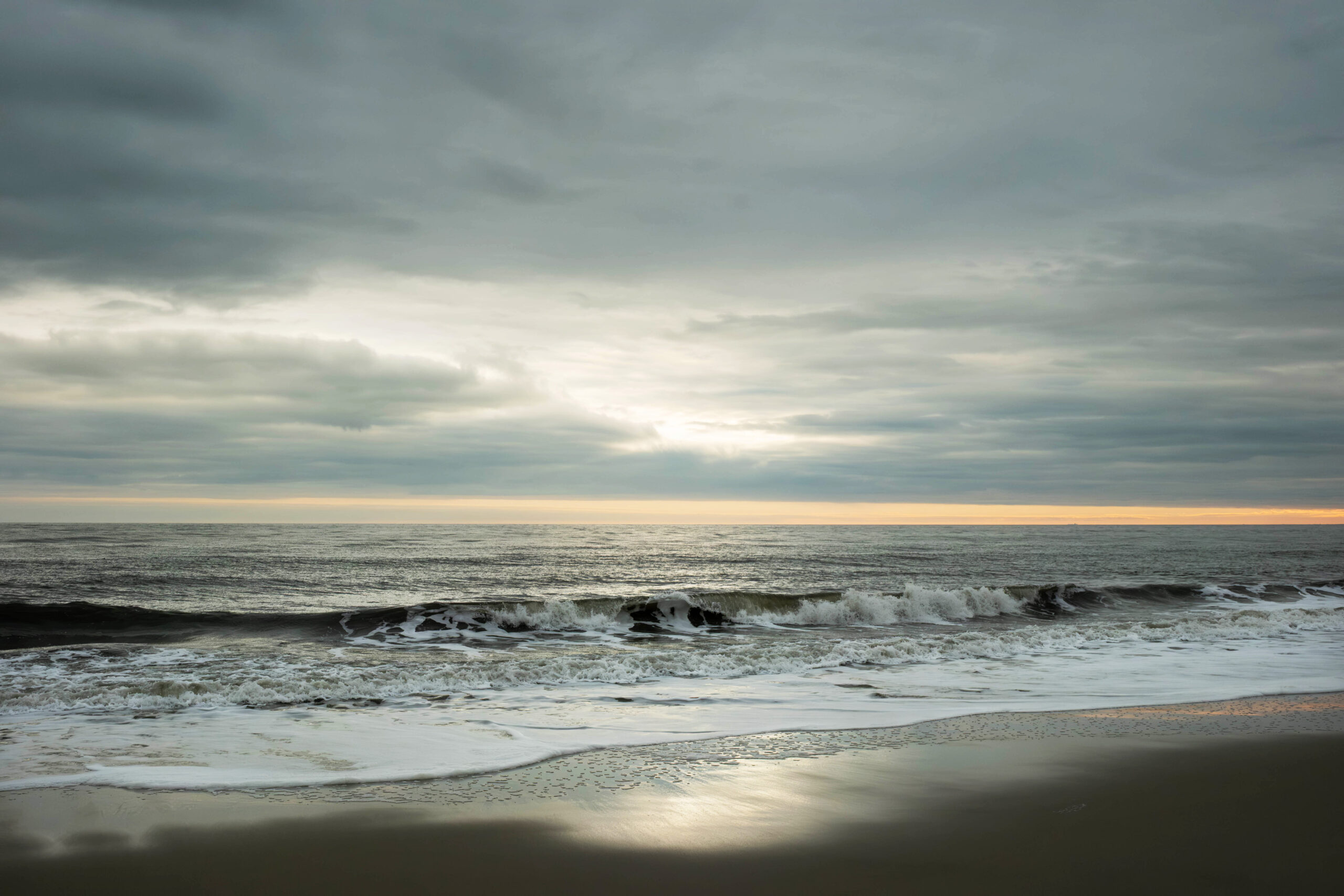 A wide view of a wave crashing in the ocean with clouds in the sky. There is a thin strip of orange light at the horizon, and sunlight is trying to break through the clouds.