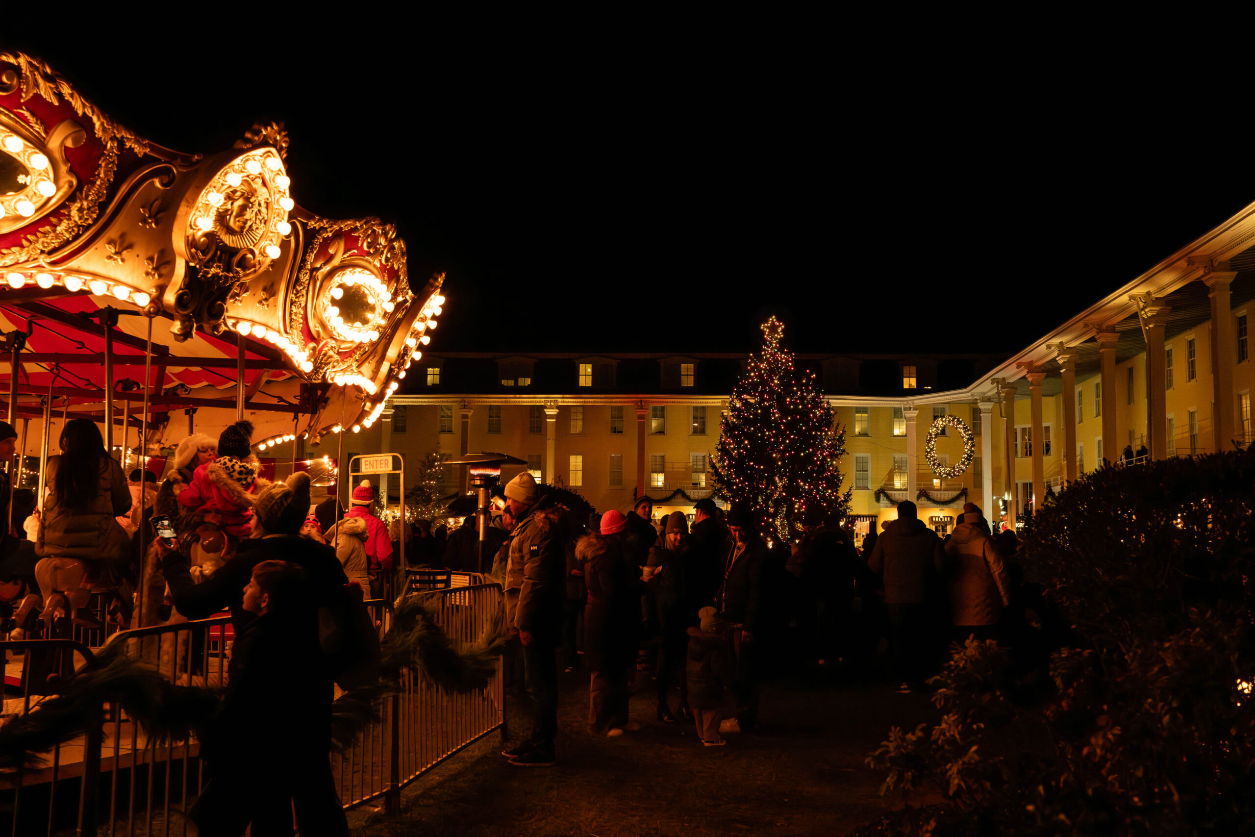 A wide view of the front lawn of Congress Hall at night. There is a Christmas tree with lights on, and part of a merry-go-round is in the foreground. There are crowds of people on the lawn.