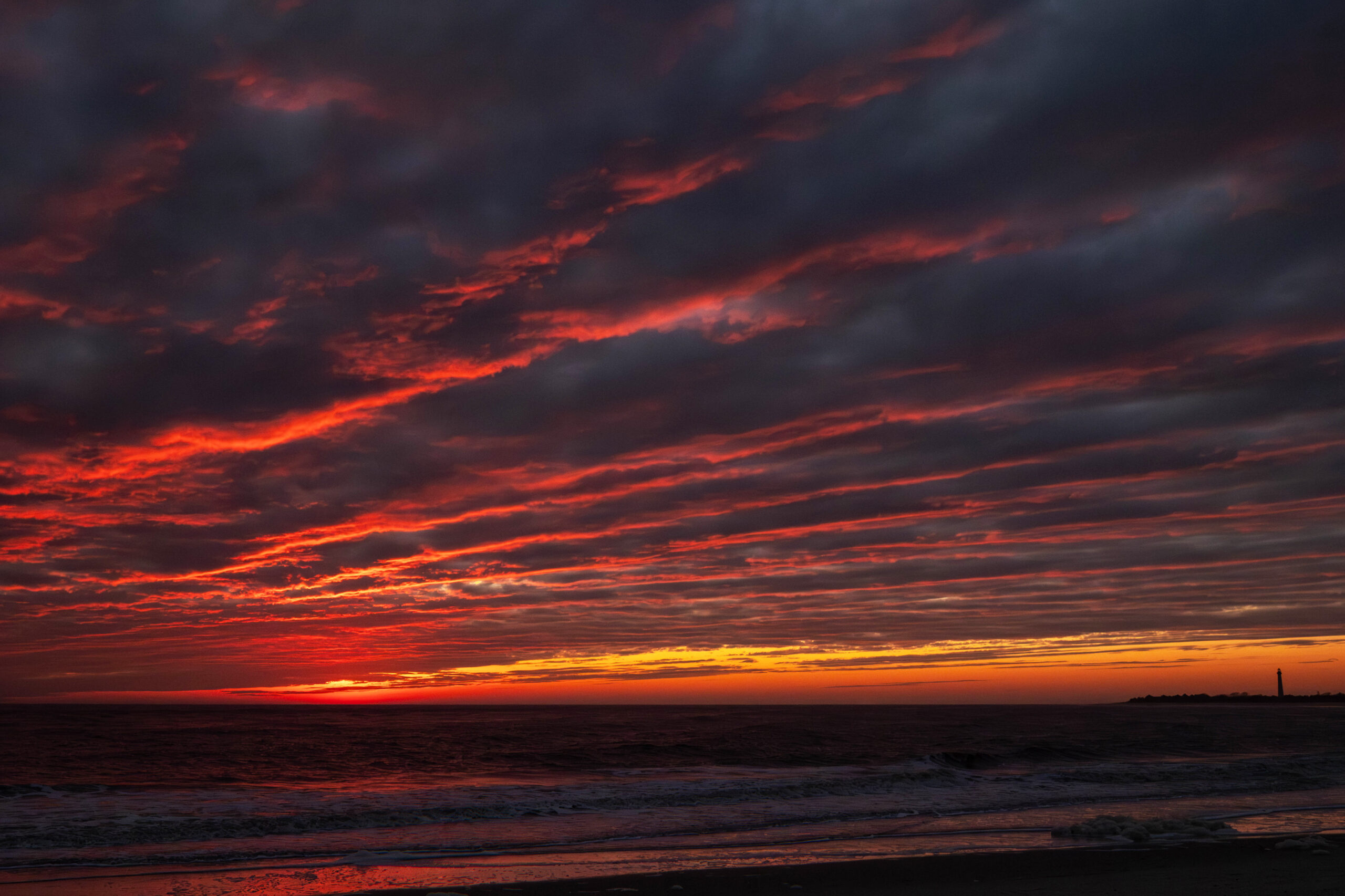 A wide view of sunset with colorful clouds in the sky. The clouds are dark purple with strips of red. The Cape May lighthouse is at the horizon line in the distance.