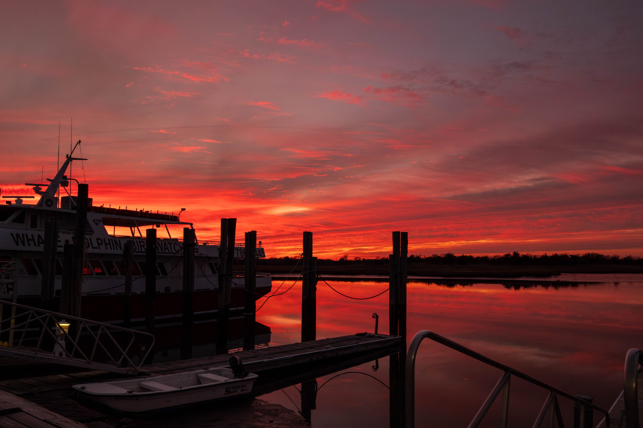 A wide view of a colorful sunset over the harbor, docks, and Cape May Whale Watcher boat. The sky has thin red, orange, and pink clouds. The colors are reflected in the water. The boat and docks are silhouetted by the sunset.